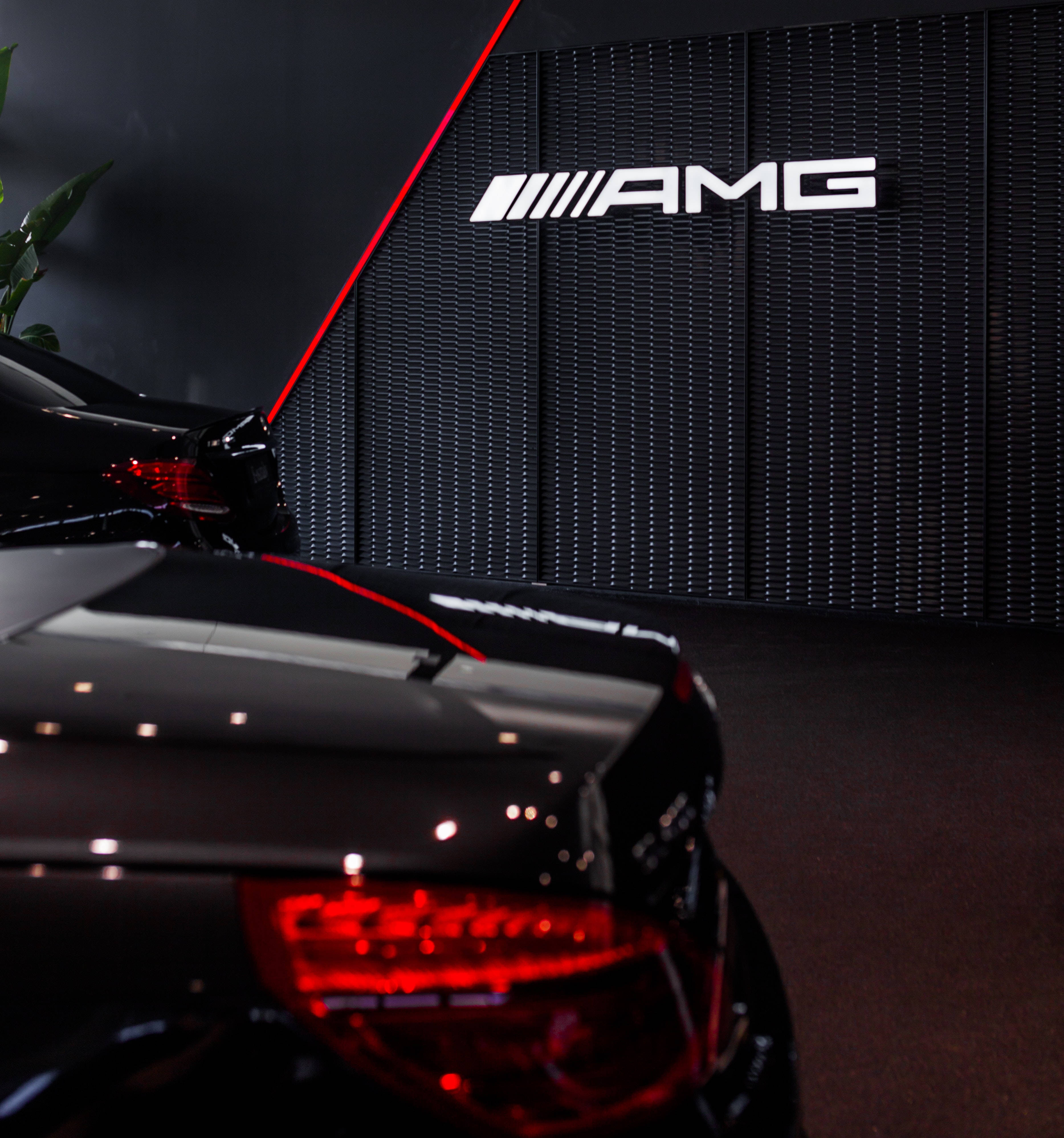 mercedes amg iPhone 桌布 壁紙 — 一輛時尚的黑色AMG跑車，帶有發光的紅色尾燈，在帶有照明AMG品牌的深色紋理牆壁前展示。