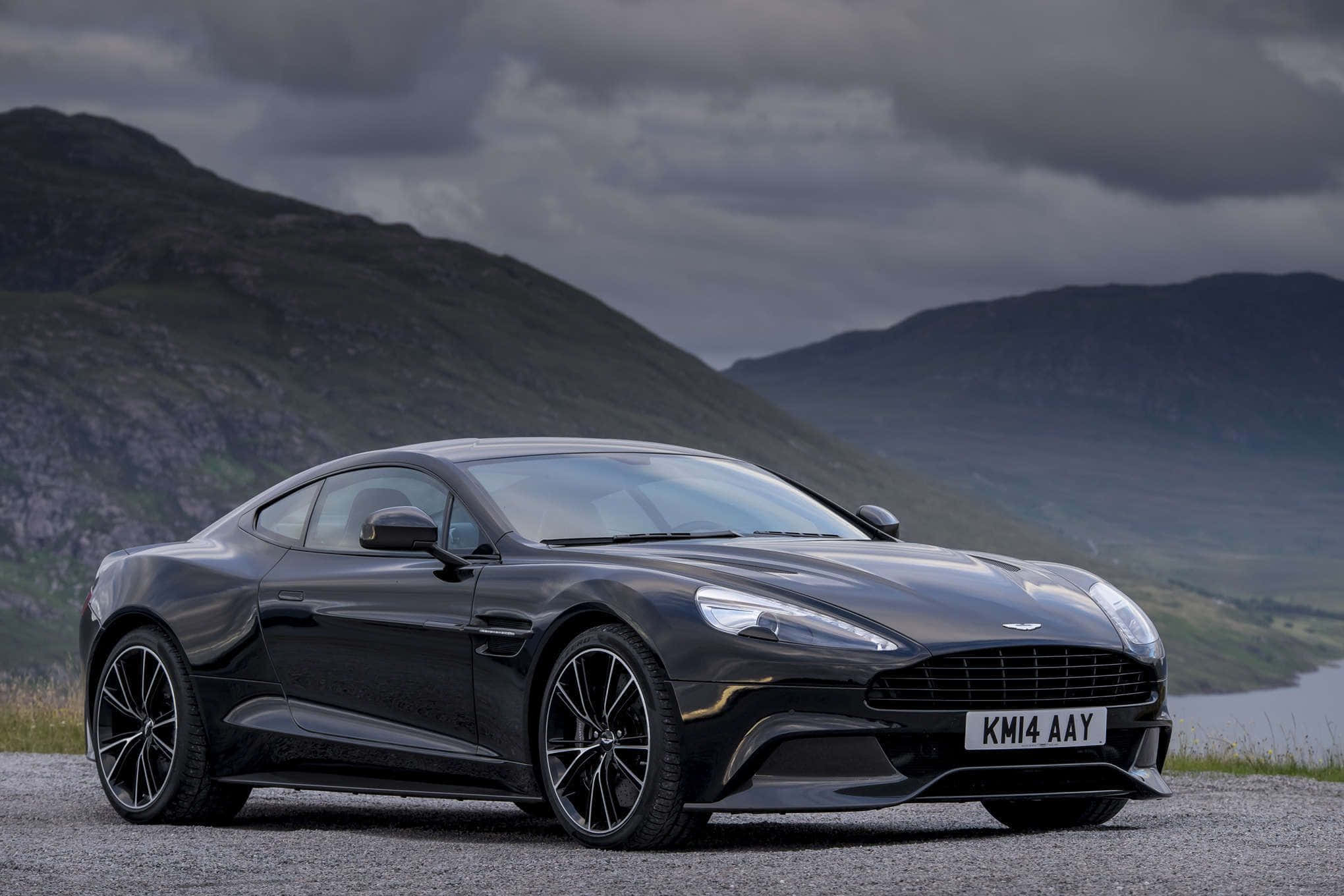 Aston Martin Vanquish 桌布 壁紙 — 一輛時尚的黑色Aston Martin跑車停泊在碎石路上，背景是戲劇性的山區地形和多雲的天空。