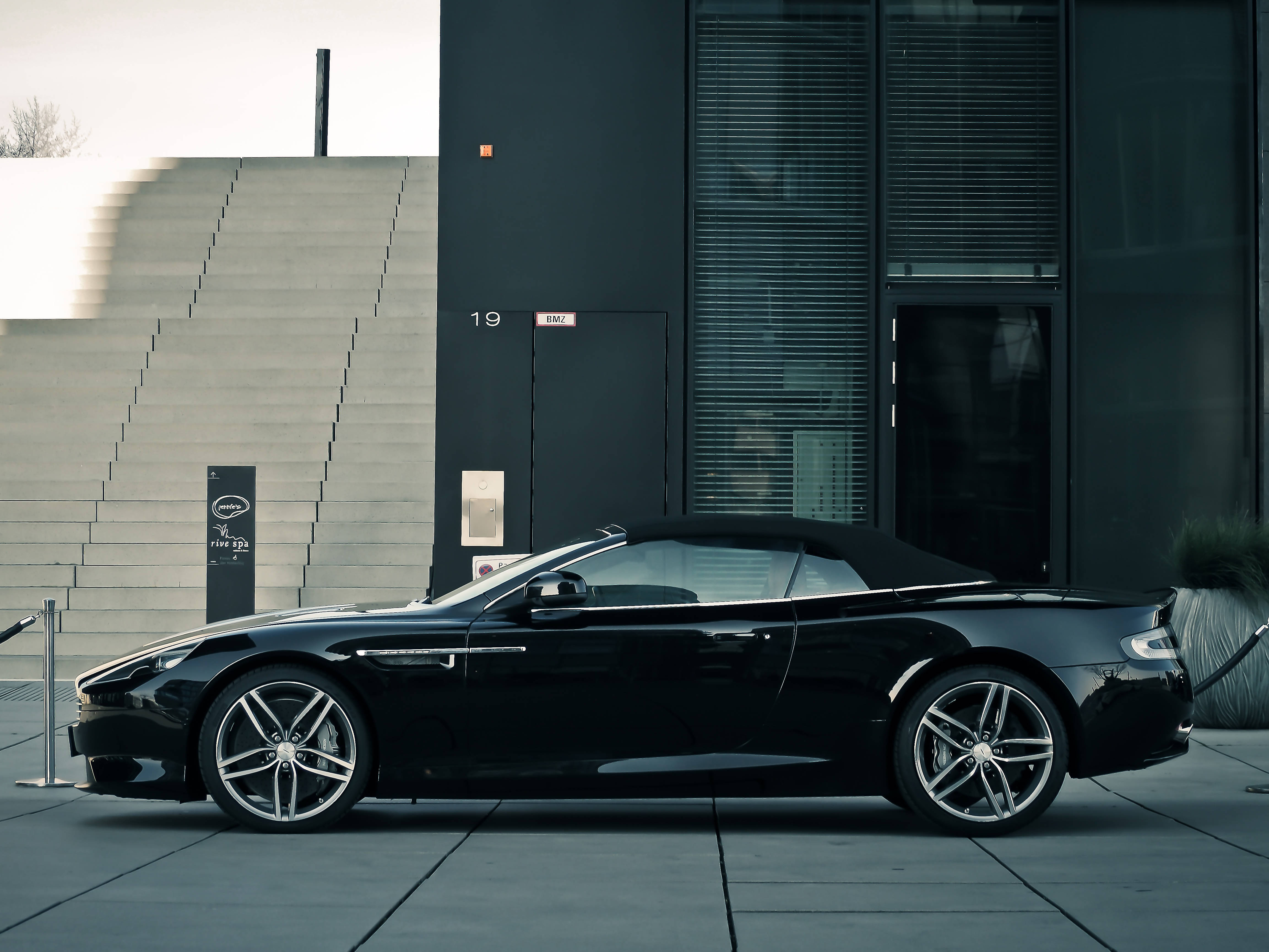 奧斯頓馬丁 桌布 壁紙 — 一輛時尚的黑色Aston Martin敞篷車停放在現代建築前，建築物有玻璃窗和混凝土台階。