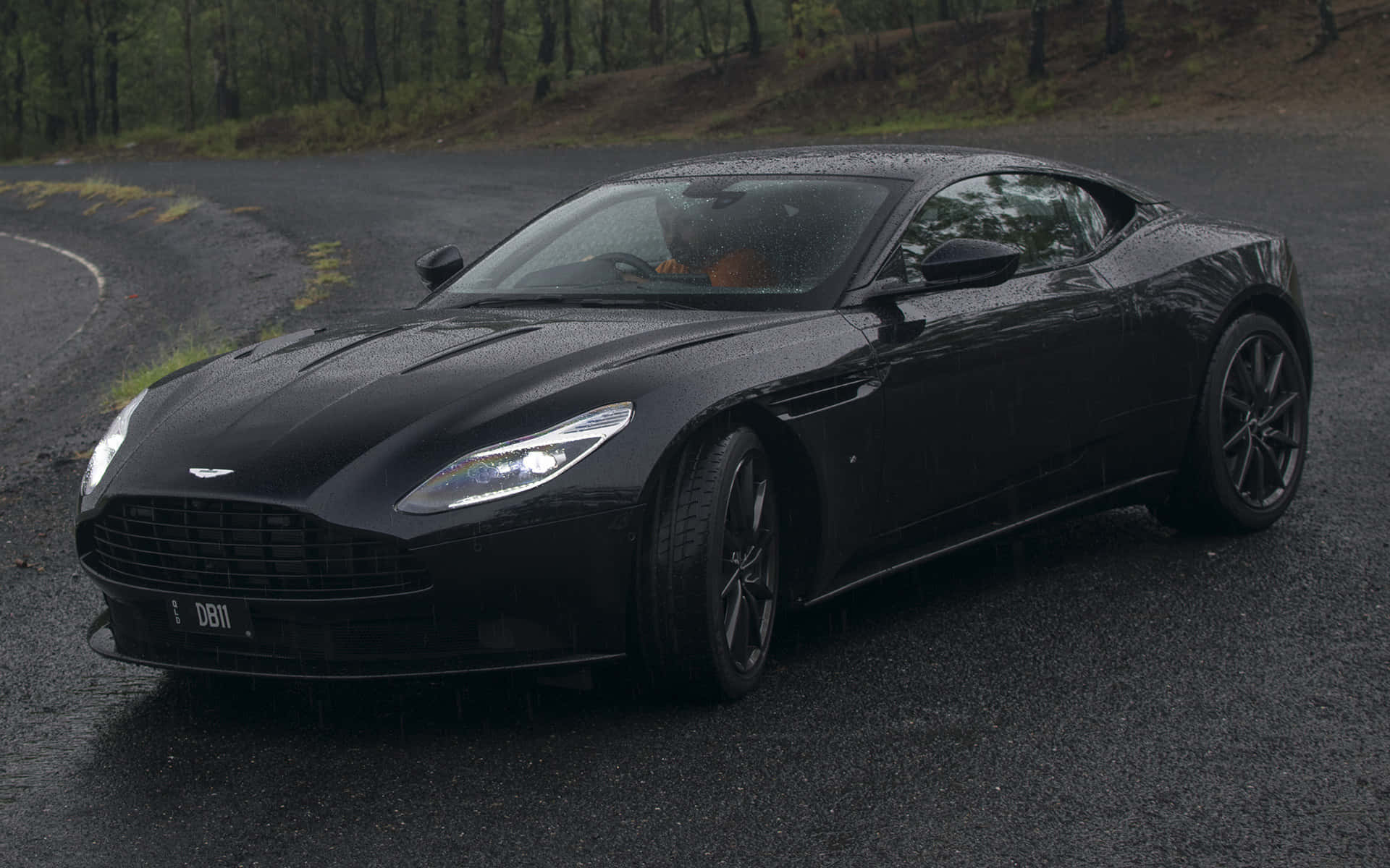 Aston Martin Db11 桌布 壁紙 — 一輛時尚的黑色Aston Martin DB11在雨中閃耀，行駛在蜿蜒的森林道路上，潮濕的瀝青反射著車頭燈。