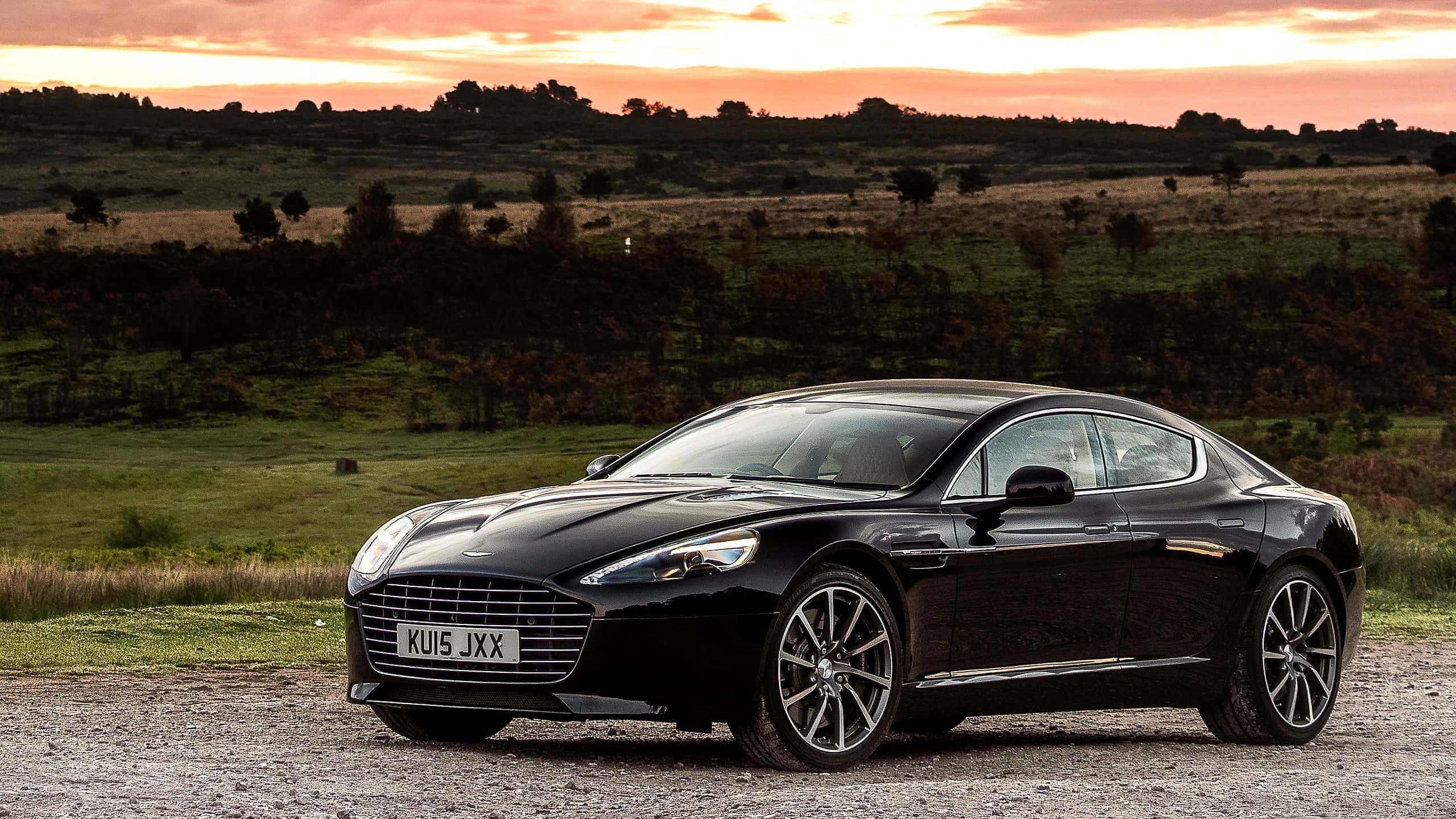 Aston Martin Rapide S 桌布 壁紙 — 一輛時尚的黑色Aston Martin跑車停在夕陽下的碎石地上，背景是連綿起伏的綠色山丘。