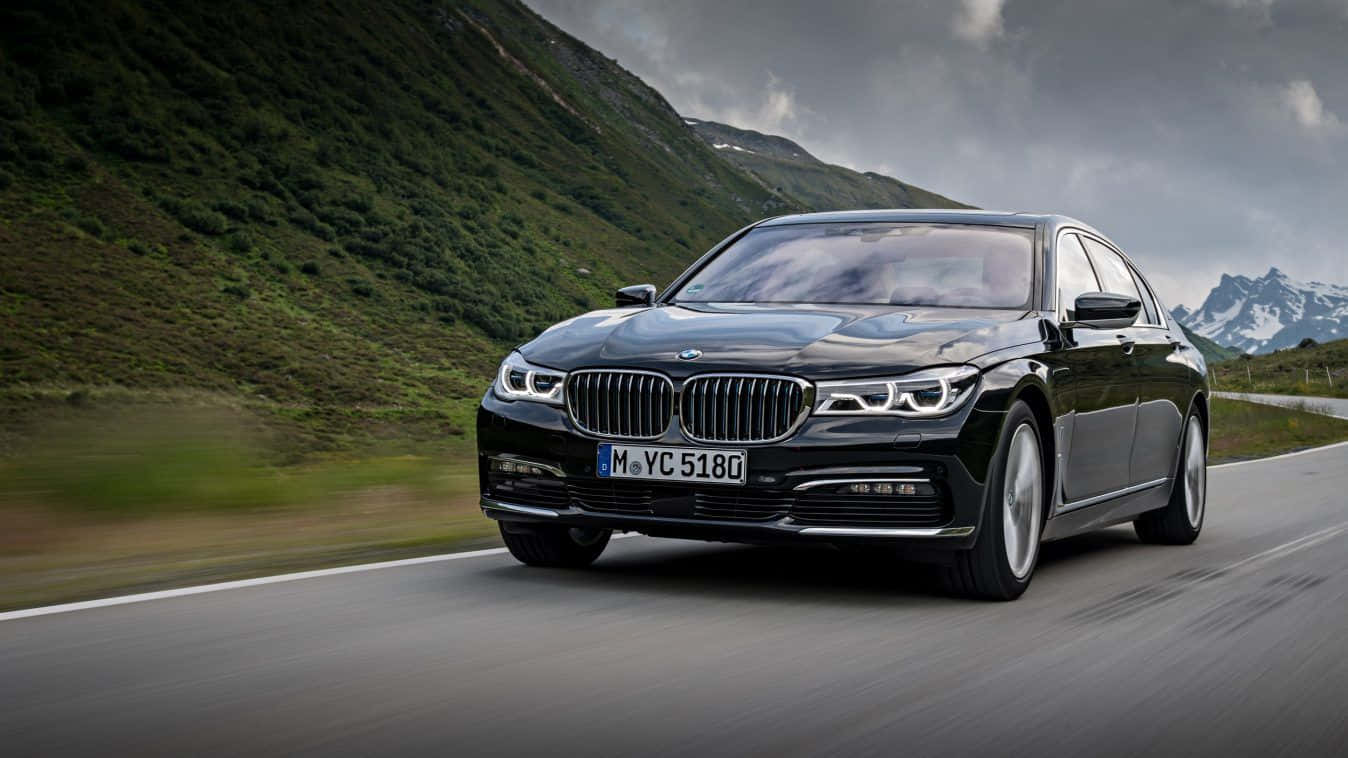 Bmw 7 Series 桌布 壁紙 — 一輛時尚的黑色BMW 7系列轎車在蜿蜒的山路上疾馳，兩旁是翠綠的山坡和積雪的山峰，天空多雲。