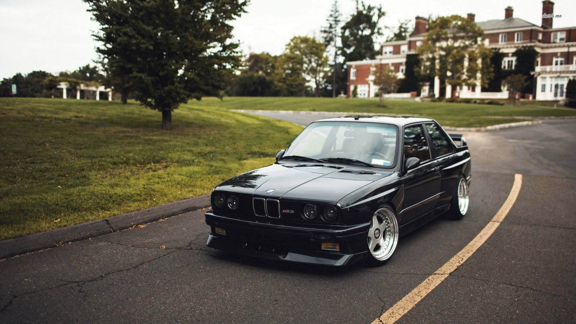 m 桌布 壁紙 — 一輛時尚的黑色BMW E30 M3跑車停泊在彎曲的瀝青路上，旁邊是修剪整齊的綠色草坪和宏偉的磚砌房屋。