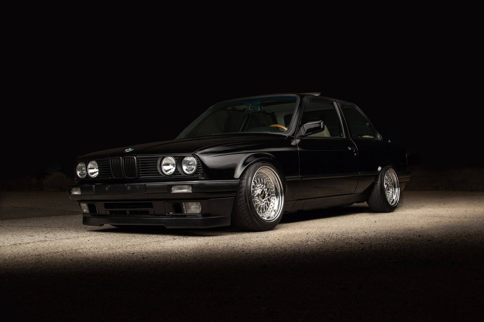 BMW 動圖 桌布 壁紙 — 一輛時尚的黑色BMW E30雙門轎跑車配備定制輪圈，在夜間於黑暗的沙漠背景下戲劇性地照亮。