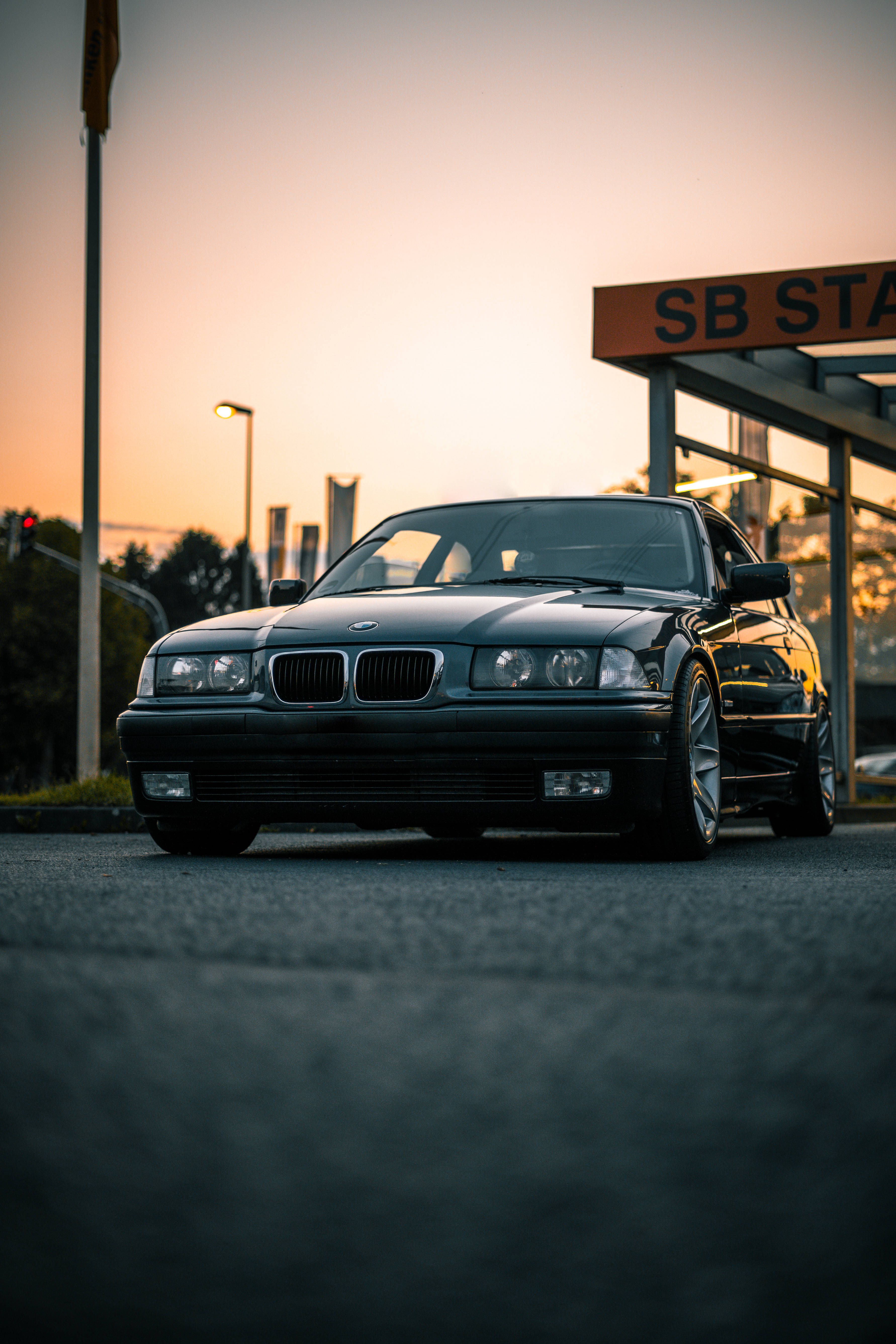 Lowrider 桌布 壁紙 — 一輛時尚的黑色BMW E38轎車在溫暖的金色日落時分停靠在加油站。