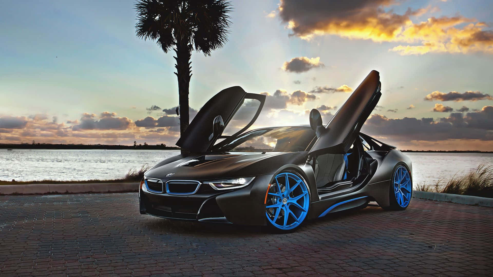 跑車 桌布 壁紙 — 一輛時尚的黑色BMW i8，配備發光藍色輪圈，停泊在日落時分水邊的磚路上，旁邊有棕櫚樹。