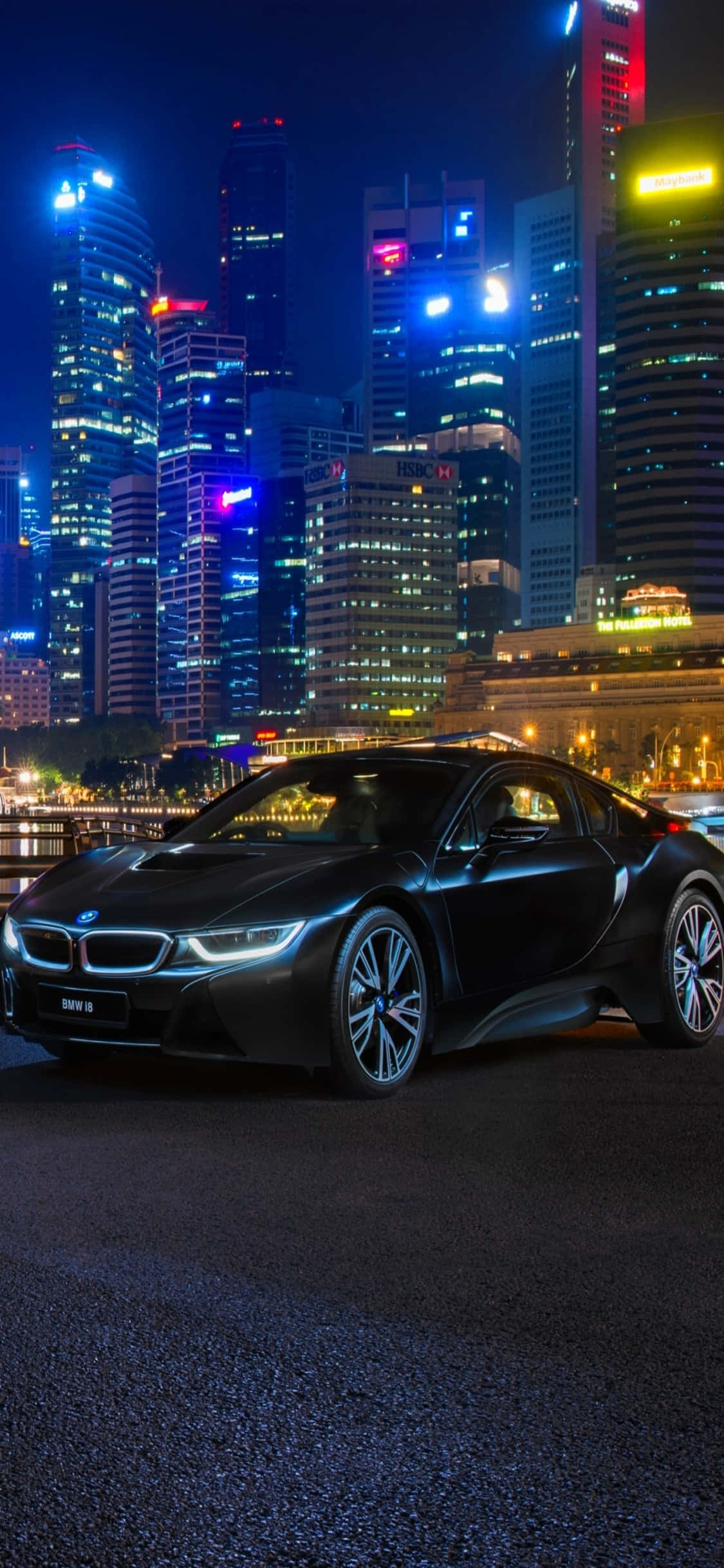 iPhone X Bmw 桌布 壁紙 — 一輛時尚的黑色BMW i8跑車在夜晚的霓虹燈下，以高聳的摩天大樓為背景閃閃發光。