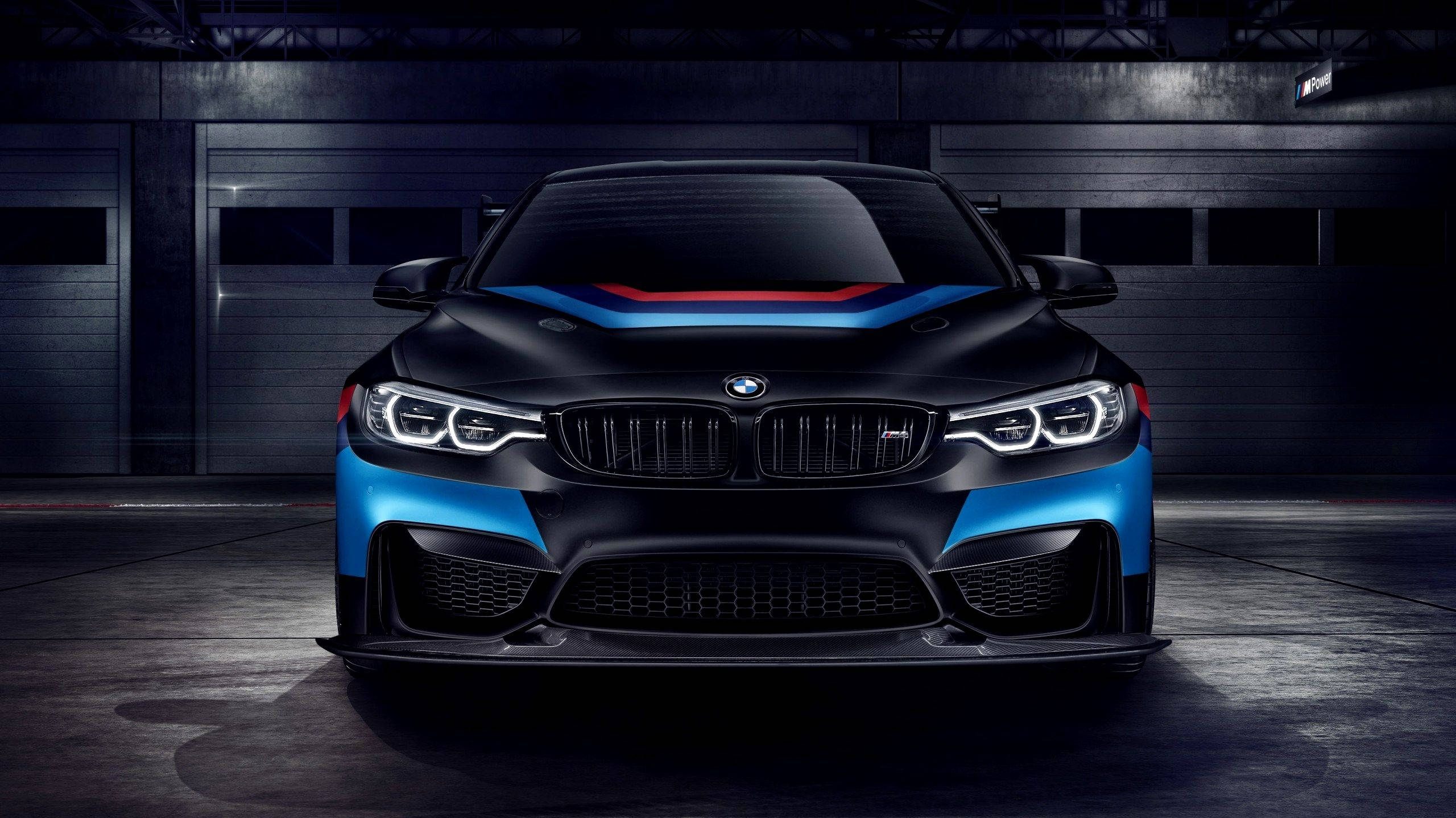 BMW 動圖 桌布 壁紙 — 一輛時尚的黑色BMW M2，帶有藍色和紅色的賽車條紋，在黑暗的工業車庫中被戲劇性地照亮。