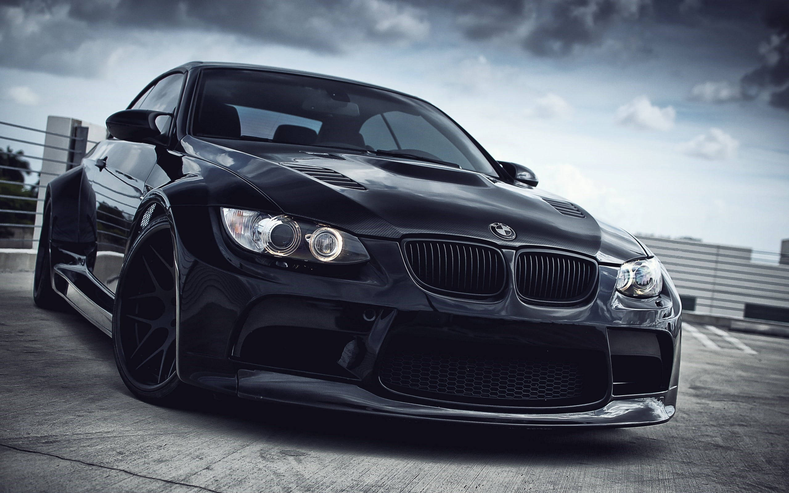 BMW 動圖 桌布 壁紙 — 一輛時尚的黑色BMW M3跑車，配備定制寬體套件和發光的頭燈，停在混凝土屋頂上，天空多雲而戲劇化。