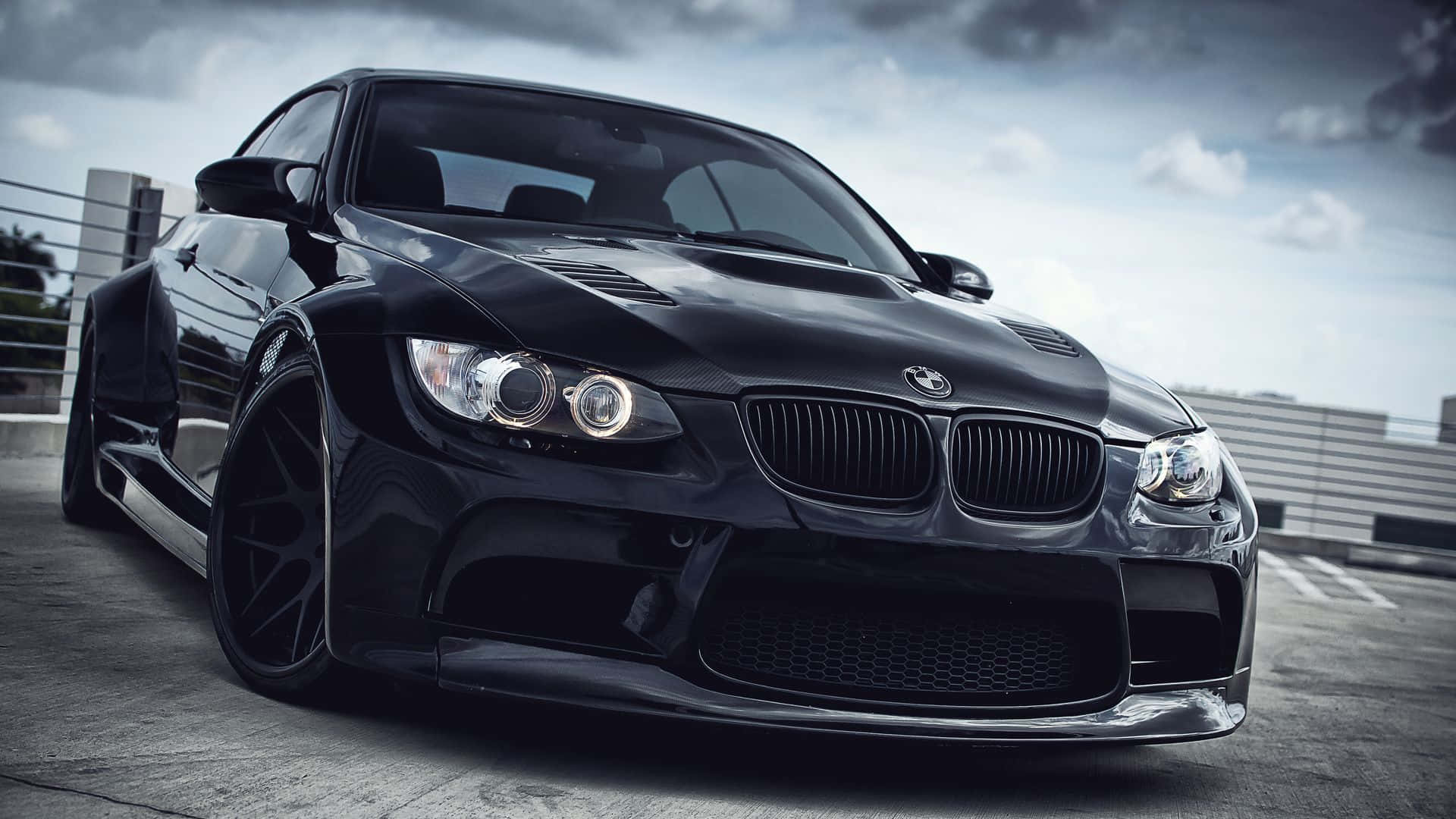 Bmw Cars 桌布 壁紙 — 一輛時尚的黑色BMW M3跑車，配備自訂寬體套件和發光頭燈，停泊在混凝土屋頂上，天空多雲。