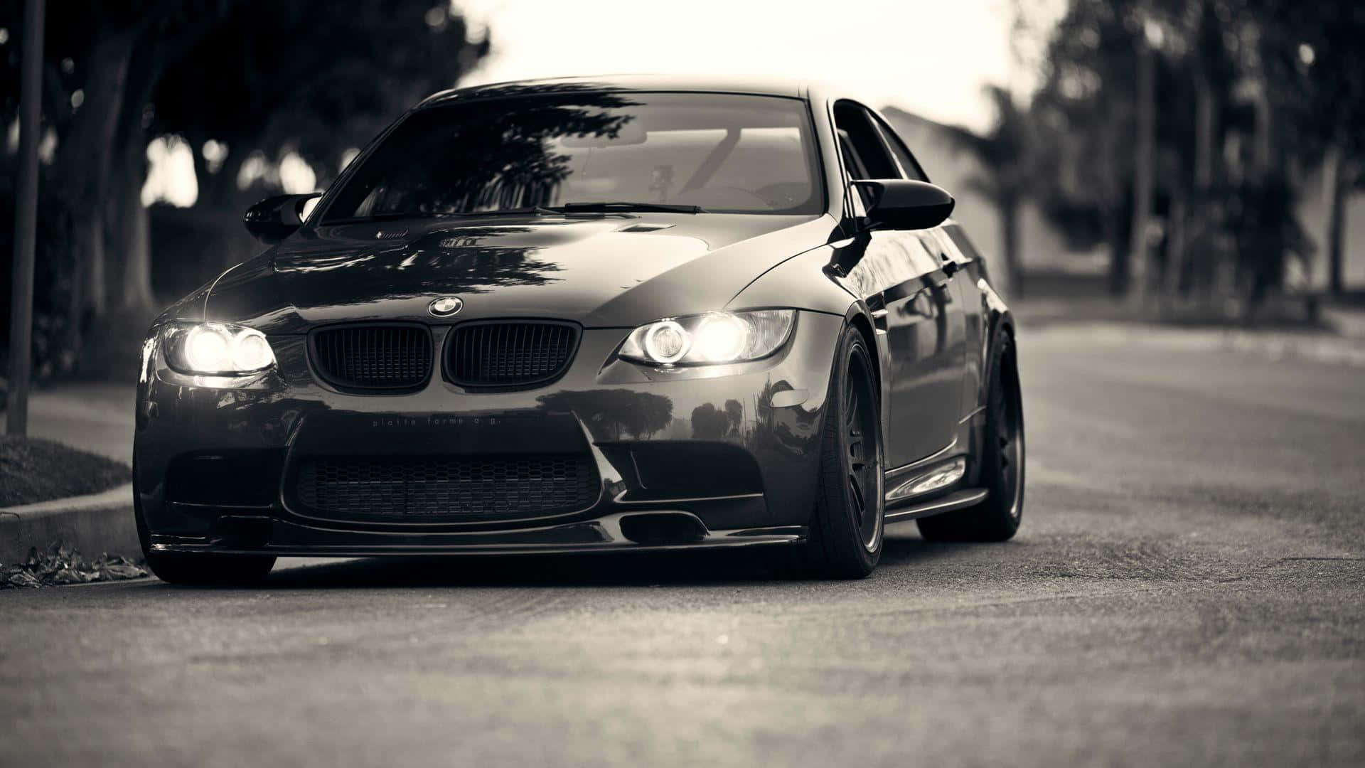 1920x1080 Bmw 桌布 壁紙 — 一輛時尚的黑色BMW M3跑車，車頭燈閃爍，以單色調停放在綠樹成蔭的街道上。