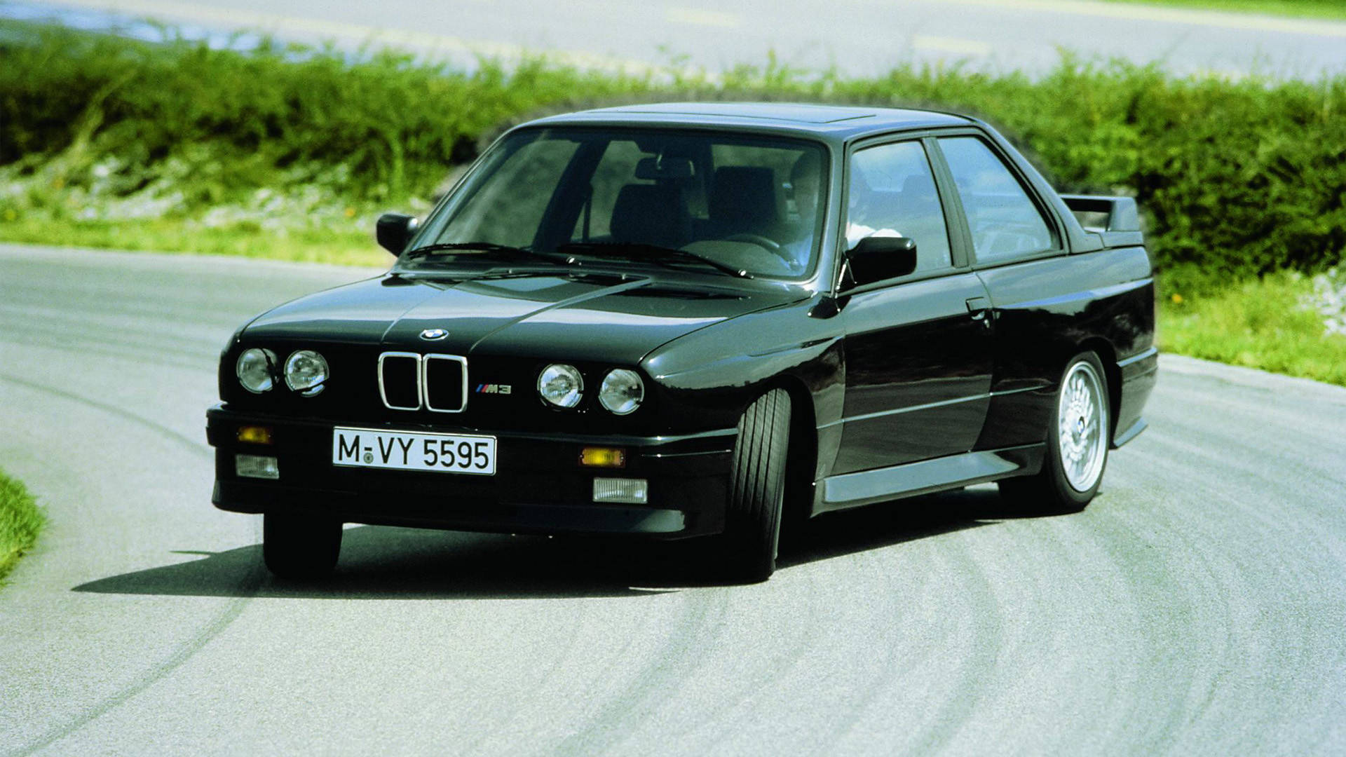 m 桌布 壁紙 — 一輛時尚的黑色BMW M3跑車在明亮的日光下行駛在一條蜿蜒的鄉村道路上，兩旁是茂密的綠色樹籬。