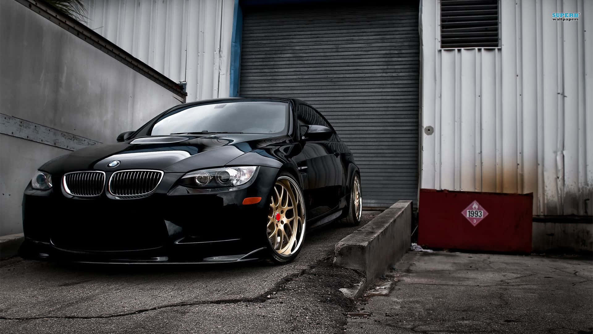 桌布 壁紙 — 一輛時尚的黑色BMW M3配金色輪圈，停在風化的工業車庫門前。