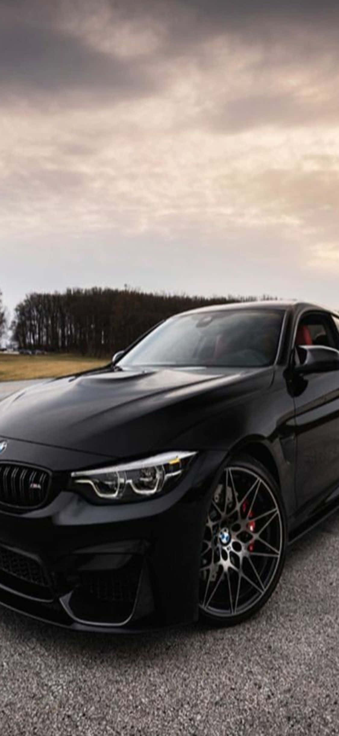 iPhone X Bmw 桌布 壁紙 — 一輛時尚的黑色BMW M4跑車停在碎石上，陰沉多雲的天空下，遠處有樹木。
