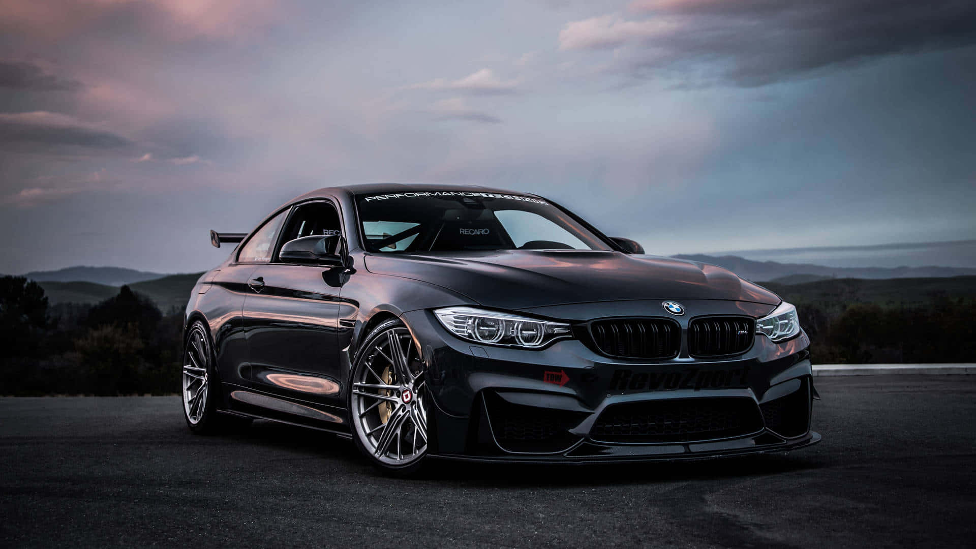 Car Images HD 桌布 壁紙 — 一輛時尚的黑色BMW M4雙門轎跑車，配備定制輪圈和後擾流板，在黃昏時分停在瀝青路上，背景是起伏的山丘。