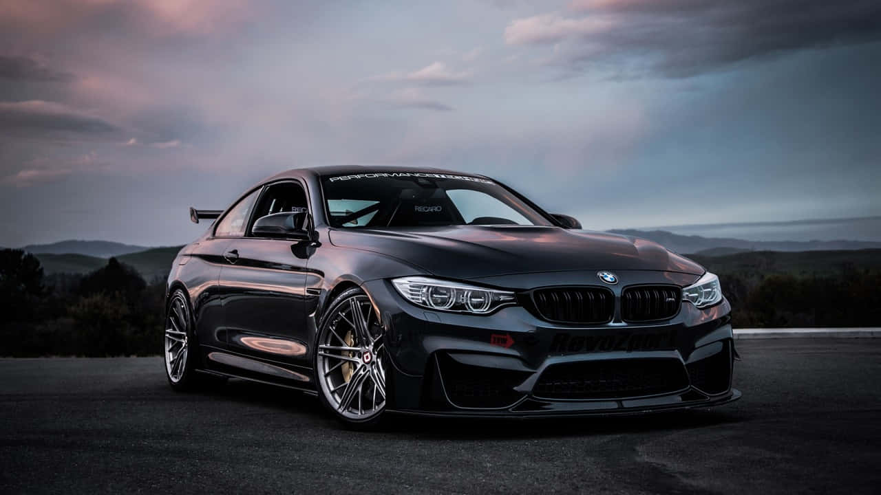720p Bmw 桌布 壁紙 — 一輛時尚的黑色BMW M4雙門轎跑車，配備定制輪轂和空氣動力學車身套件，在黃昏時分停在一條道路上，背景是起伏的丘陵。