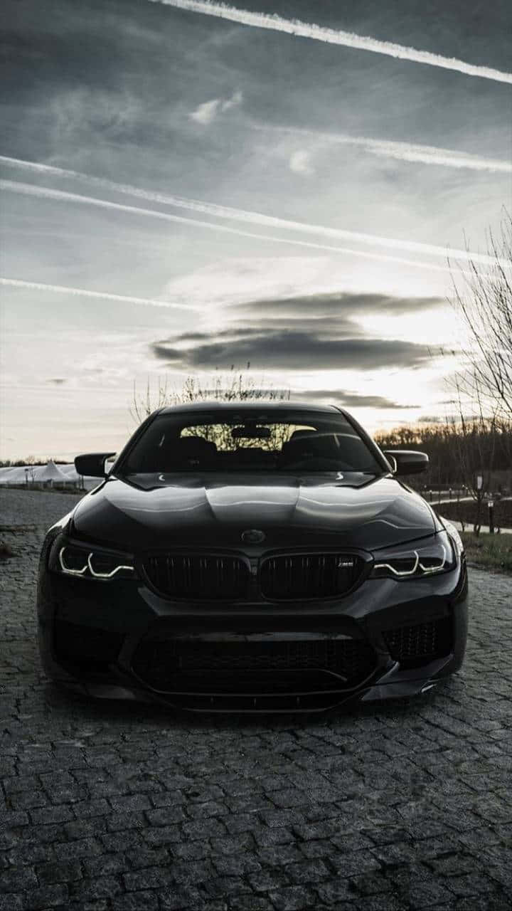 Bmw iPhone 桌布 壁紙 — 一輛時尚的黑色BMW M5停在鵝卵石路上，天空戲劇性，有凝結尾跡和柔和夕陽餘暉。