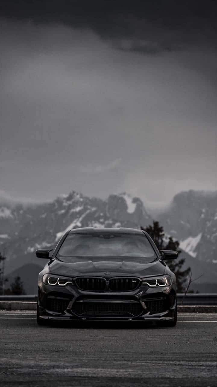 Bmw iPhone 桌布 壁紙 — 一輛時尚的黑色BMW M5在風暴般的灰色天空下，面向山路上，背景是白雪覆蓋的山峰。