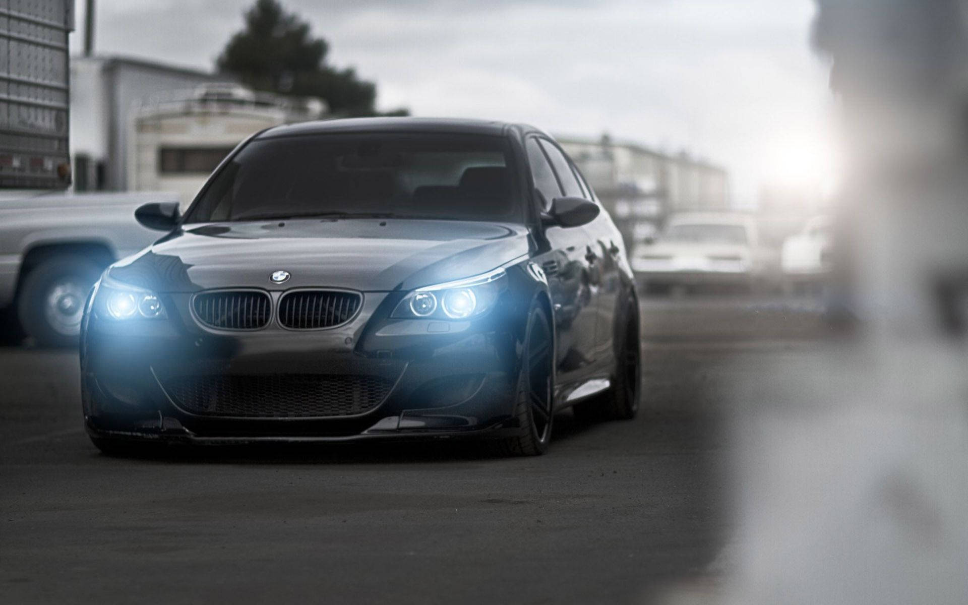 m 桌布 壁紙 — 一輛時尚的黑色BMW M5跑車，配備發光的車頭燈，在多雲的天空下主宰著一個粗糙的城市地段。