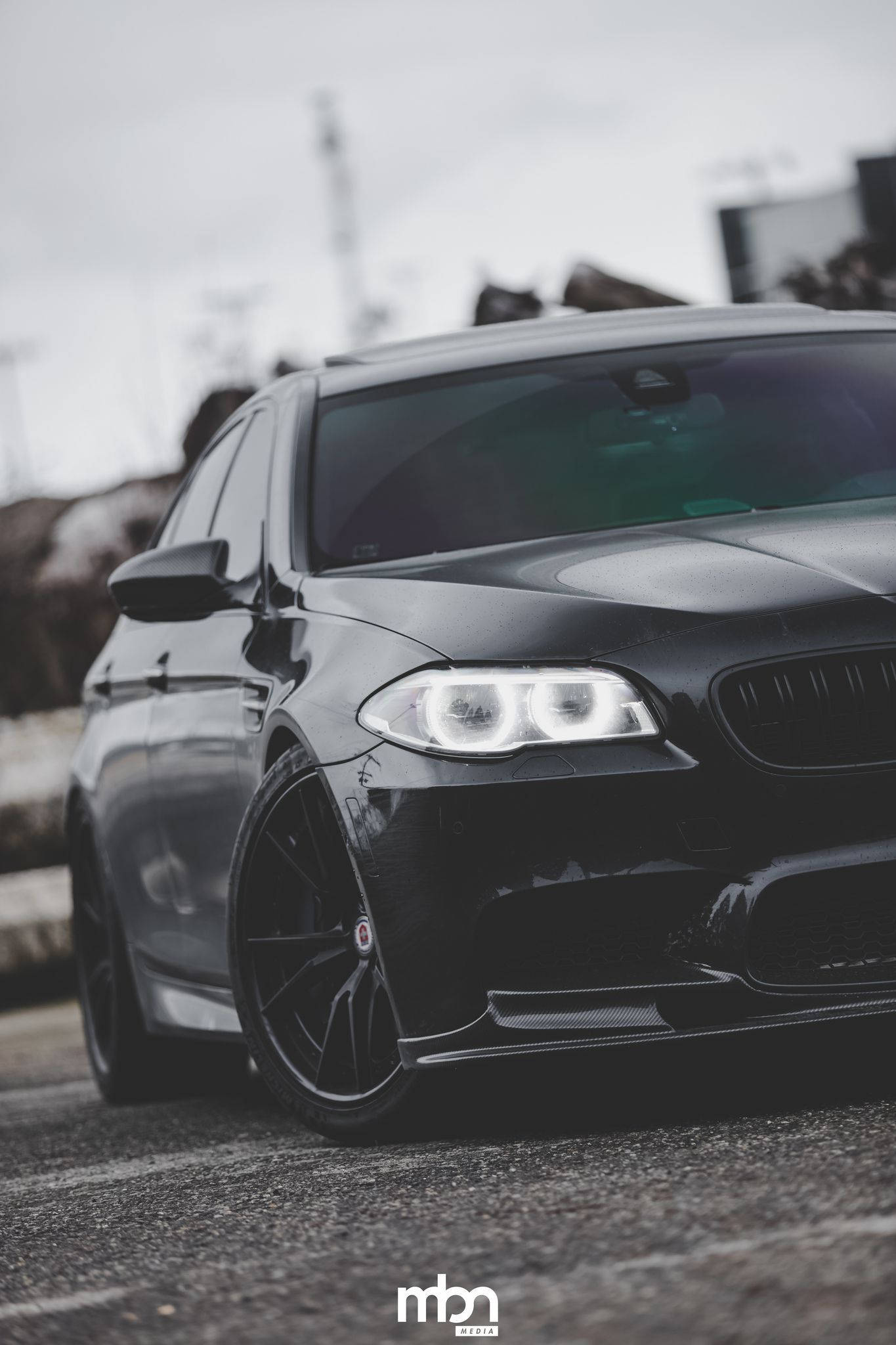 X 桌布 壁紙 — 一輛時尚的黑色BMW M5跑車，配備發光頭燈，停泊在粗糙的瀝青路面上，天空多雲。