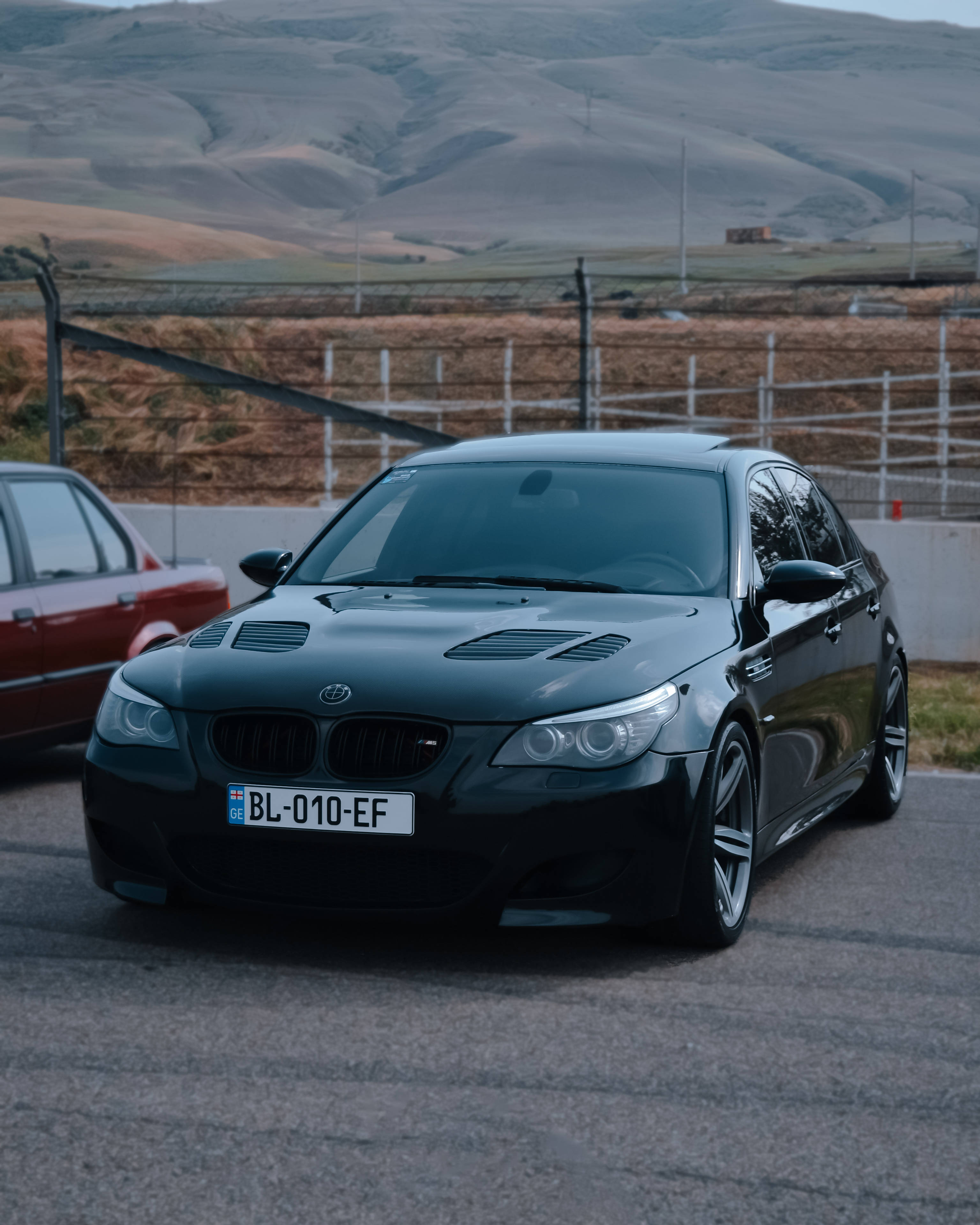 Racing 桌布 壁紙 — 一輛時尚的黑色BMW M5運動轎車停在柏油路上，背景是起伏的丘陵和圍欄。