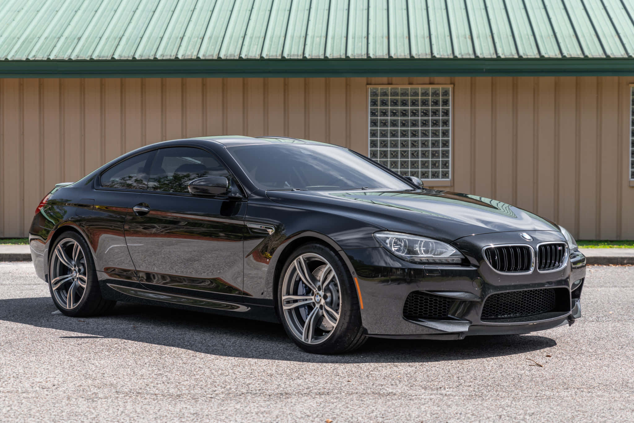 Bmw M6 桌布 壁紙 — 一輛時尚的黑色BMW M6轎跑車在明亮的陽光下閃閃發光，停在柏油路面上，前方是米色建築和綠色金屬屋頂。