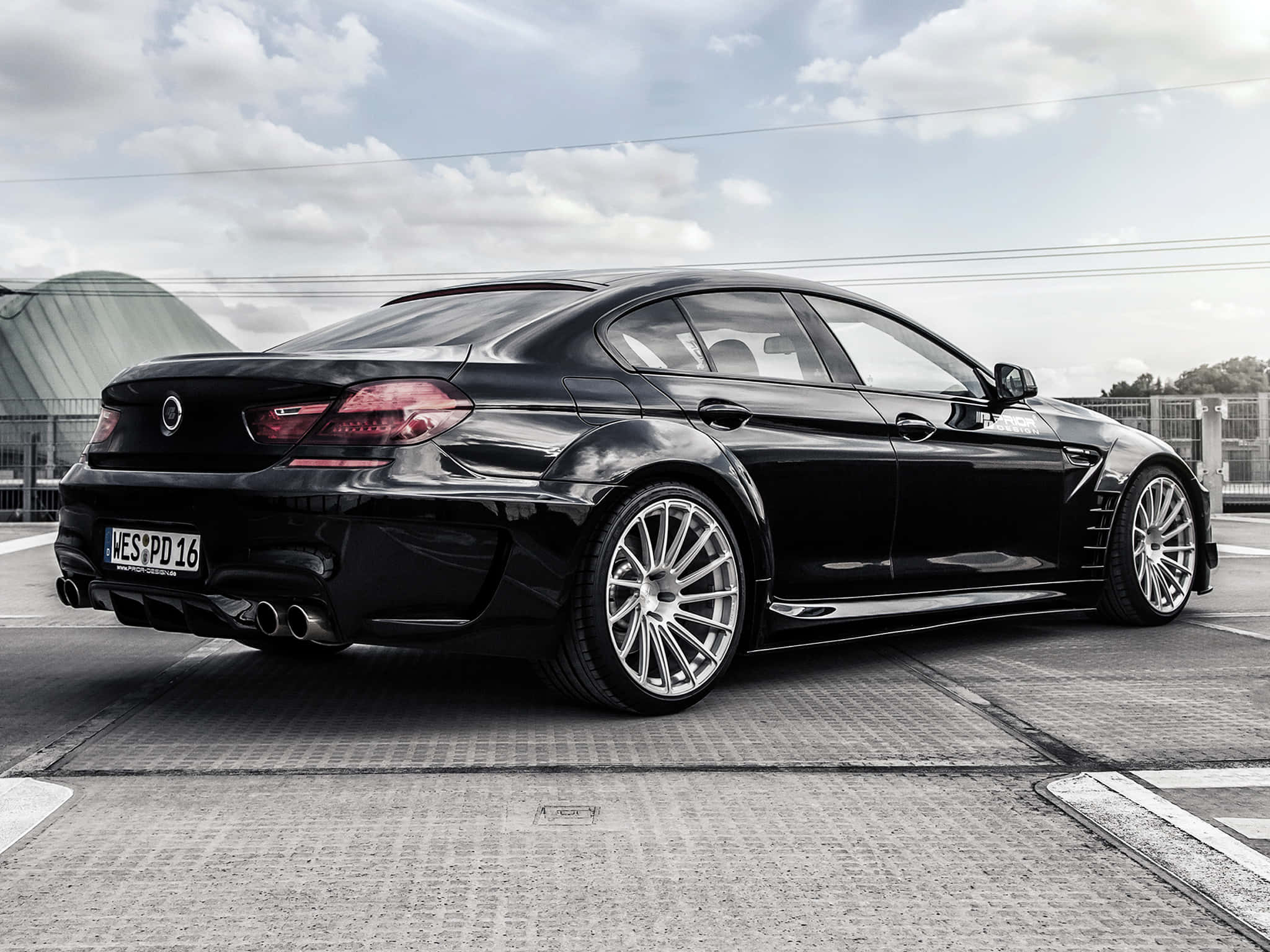 Bmw M6 桌布 壁紙 — 一輛時尚的黑色BMW M6雙門轎跑車，配備定制銀色輪圈，停泊在紋理路面上，天空多雲。
