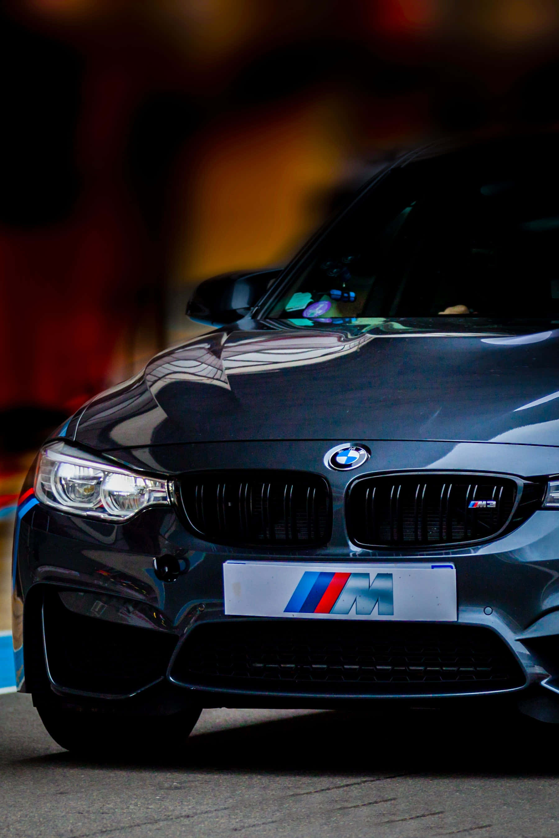 Bmw Android 桌布 壁紙 — 一輛時尚的黑色BMW M汽車，帶有發光的車頭燈和模糊的溫暖背景。