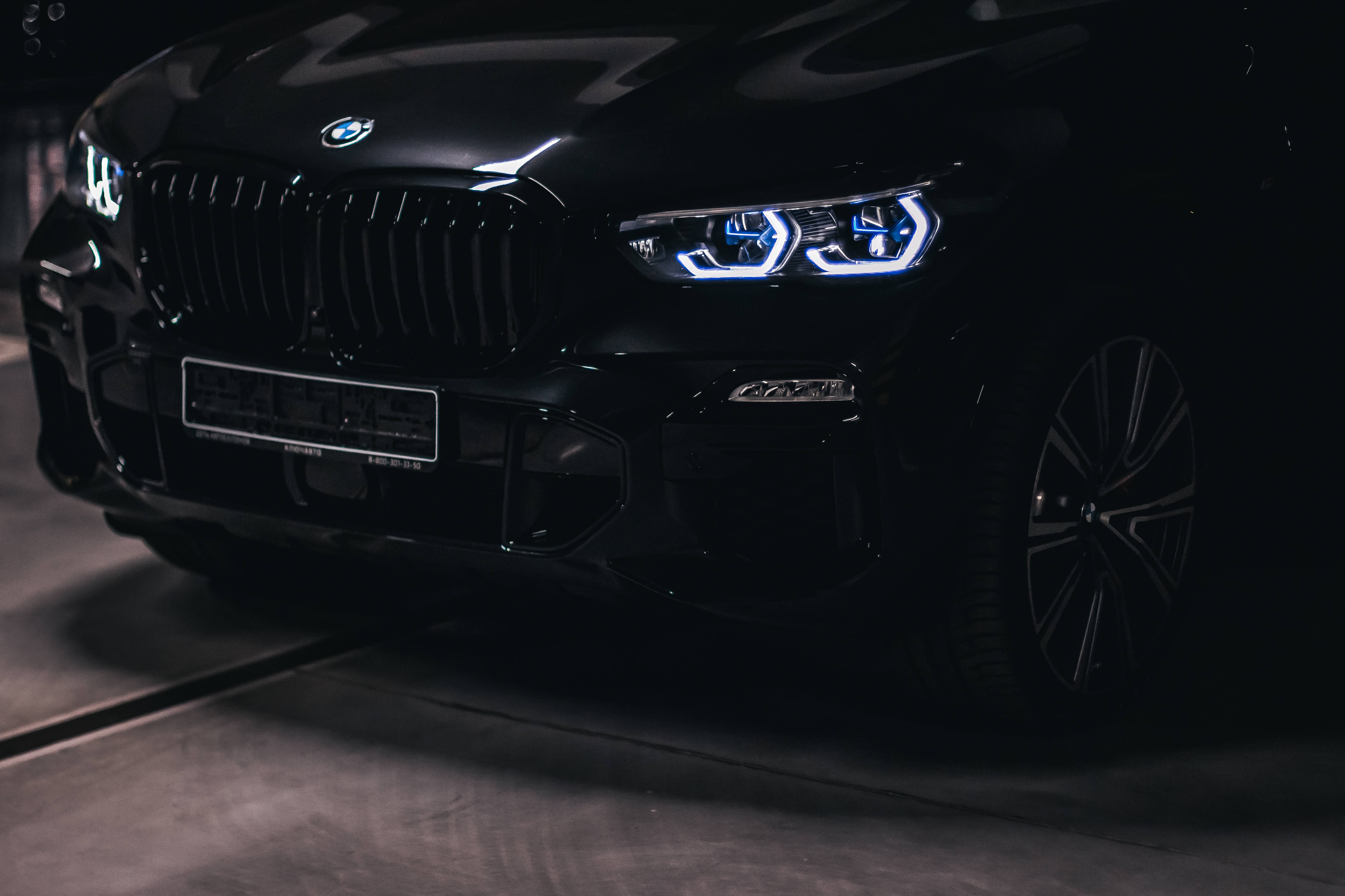 Black Bmw 桌布 壁紙 — 一輛時尚的黑色BMW SUV在昏暗的車庫中發出銳利的藍色車頭燈光，在混凝土上投射出戲劇性的陰影。