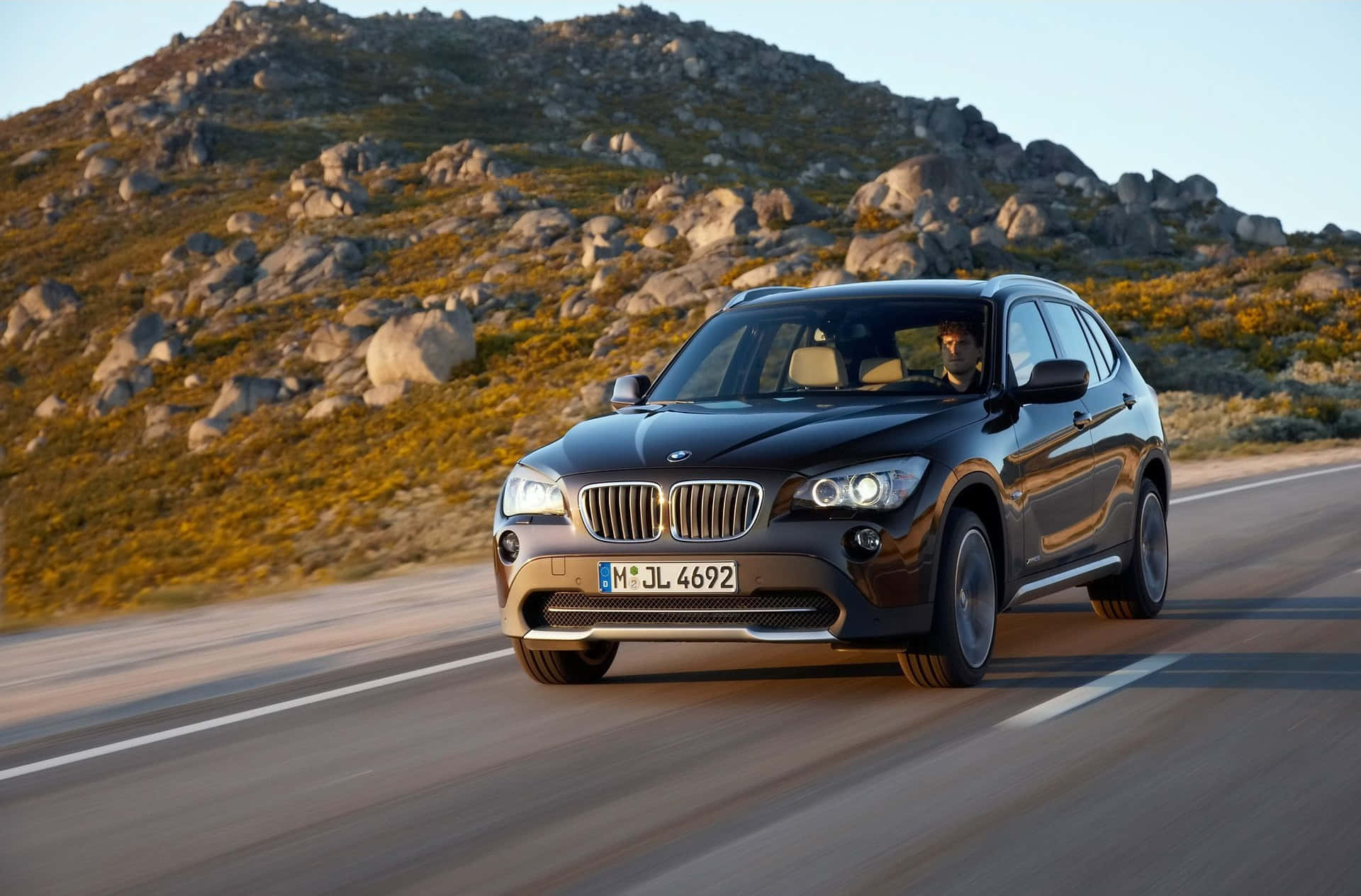 Bmw X1 桌布 壁紙 — 一輛時尚的黑色BMW X1 SUV在晴朗的天空下行駛在蜿蜒的山路上，周圍是岩石地形和金色灌木。