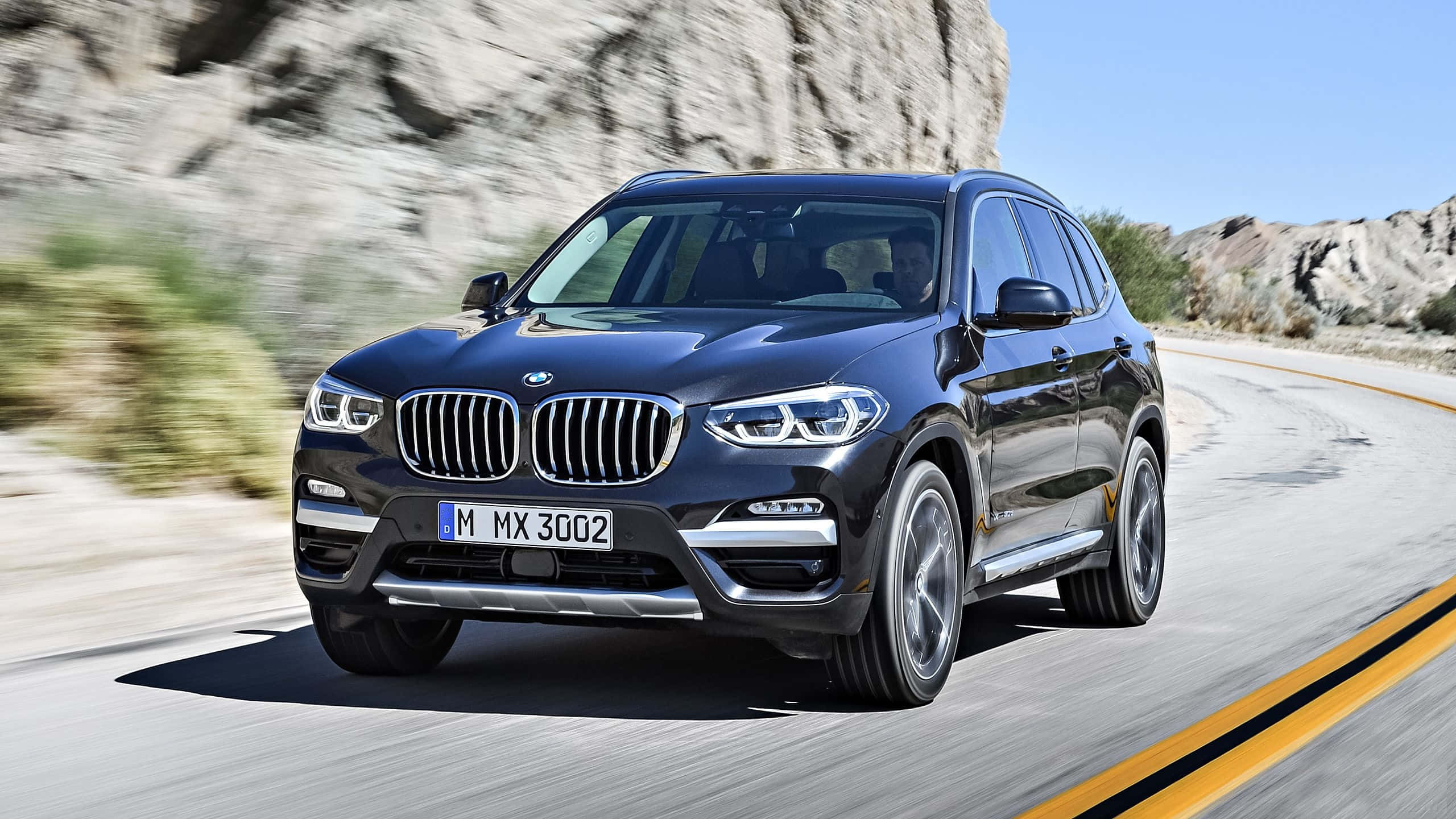 Bmw X3 桌布 壁紙 — 一輛時尚的黑色BMW X3 SUV在晴朗藍天下，沿著蜿蜒的沙漠道路疾馳，兩旁是岩石峭壁和稀疏的綠色植物。