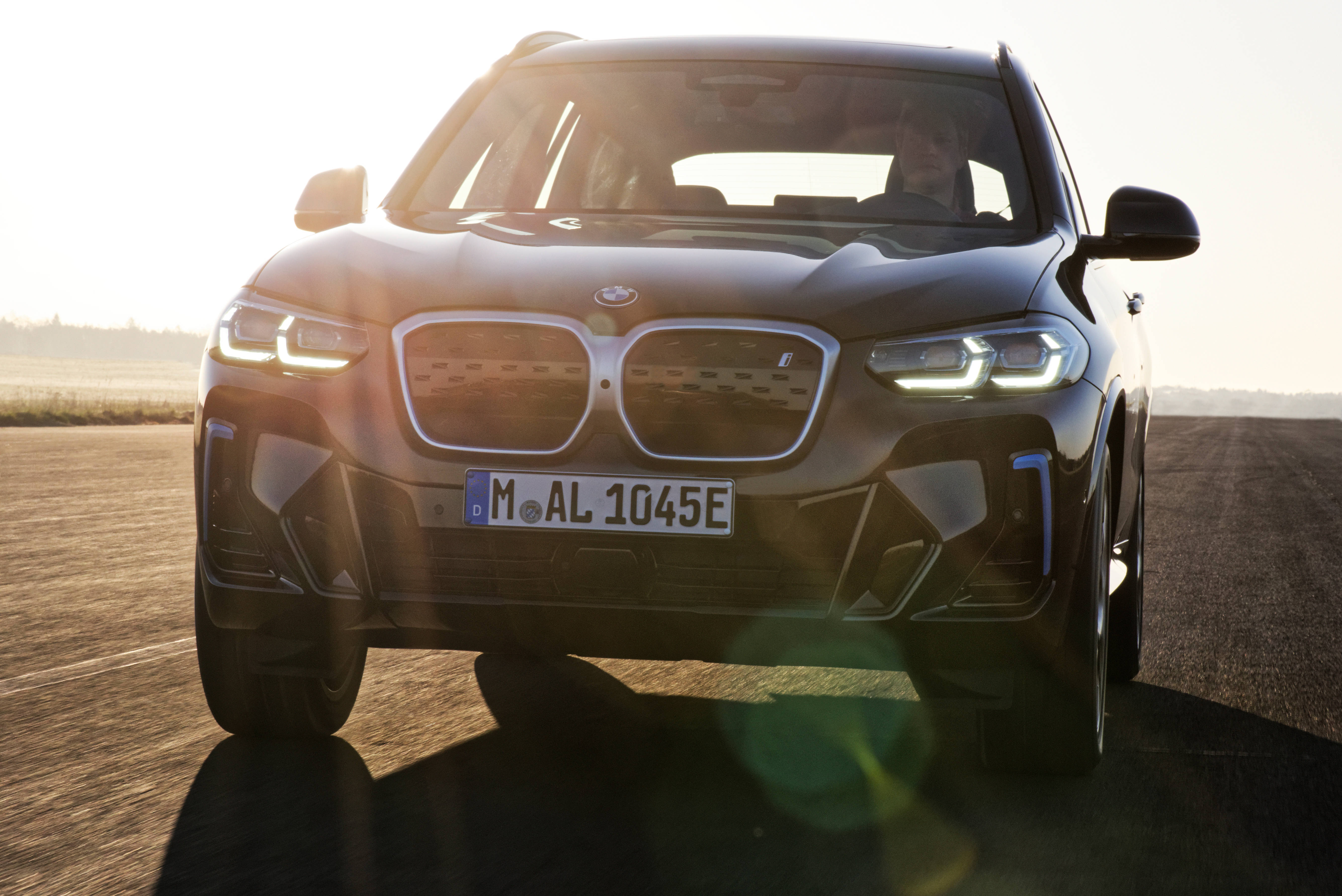 Black Bmw 桌布 壁紙 — 一輛時尚的黑色BMW X3 SUV在陽光明媚的道路上向前行駛，車頭燈閃爍並帶有鏡頭光暈。