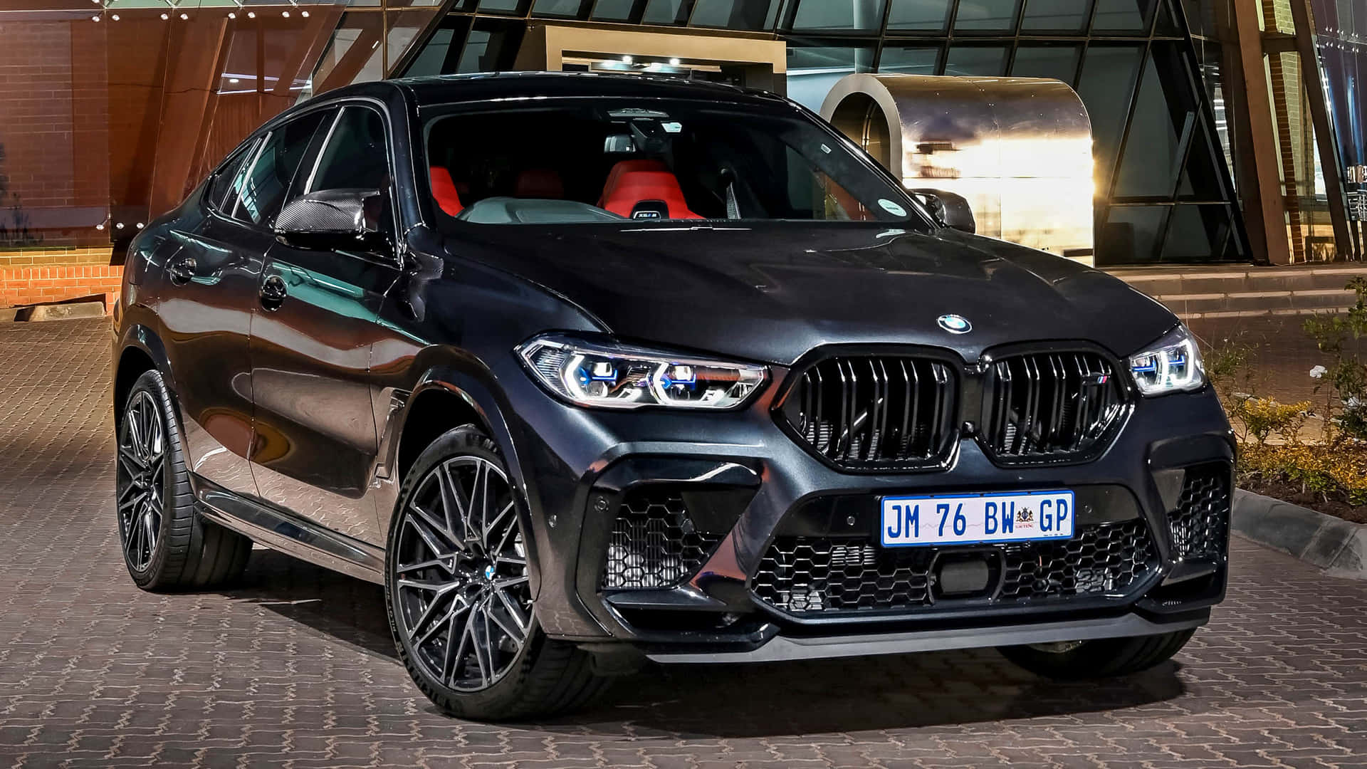 Bmw X6 M 桌布 壁紙 — 一輛時尚的黑色BMW X6 M在夜間停放在圖案人行道上，背景是現代建築。