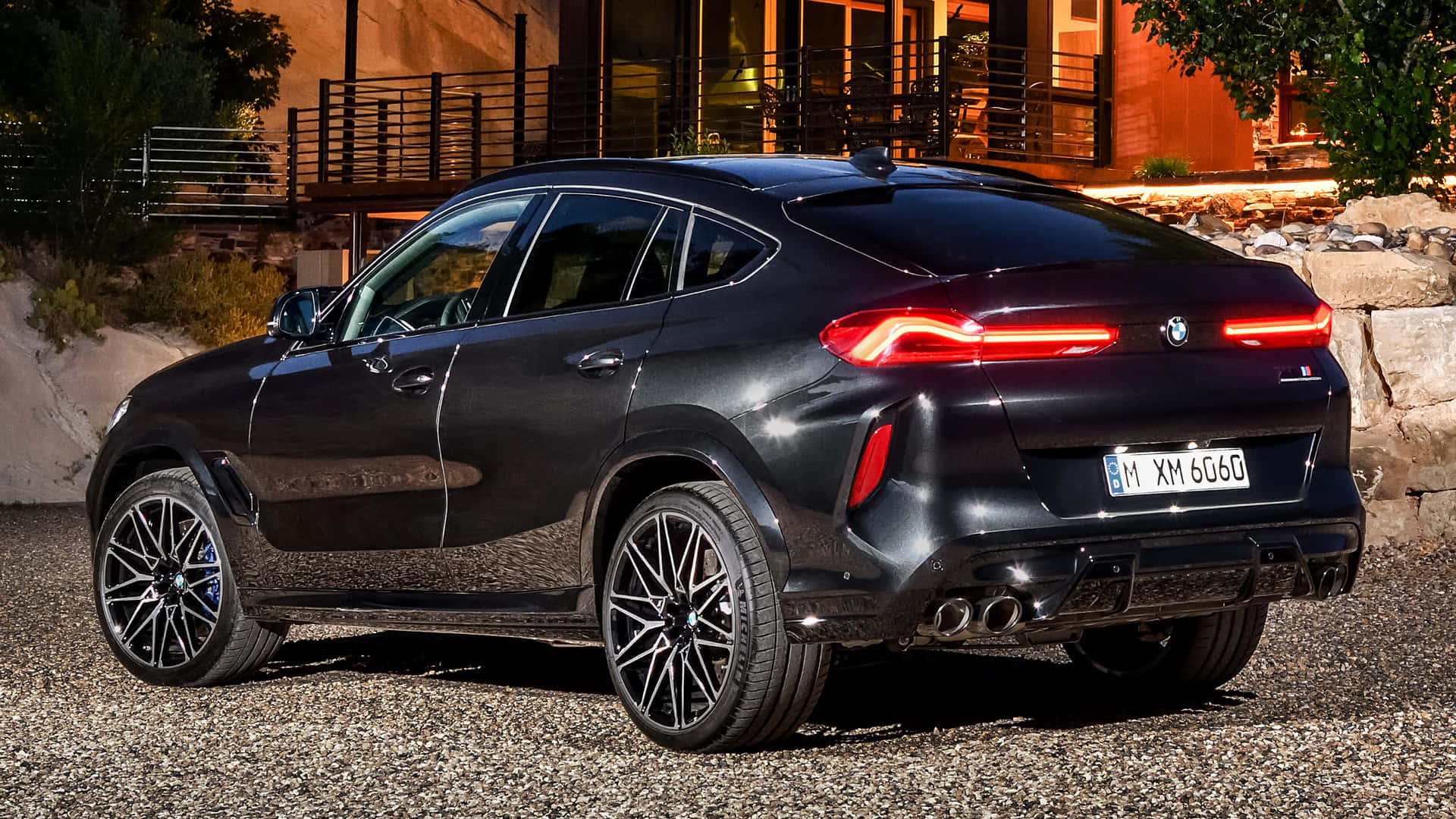 BMW X6 M 桌布 壁紙 — 一輛時尚的黑色BMW X6M停在黃昏的碎石路上，尾燈閃爍，背景是一座現代住宅。