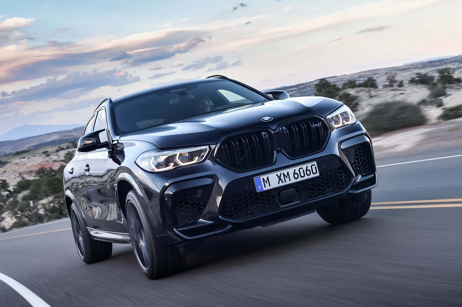 BMW X6 M 桌布 壁紙 — 一輛時尚的黑色BMW X6M在沙漠高速公路上疾馳，天空柔和暮色，雲朵零星散落。