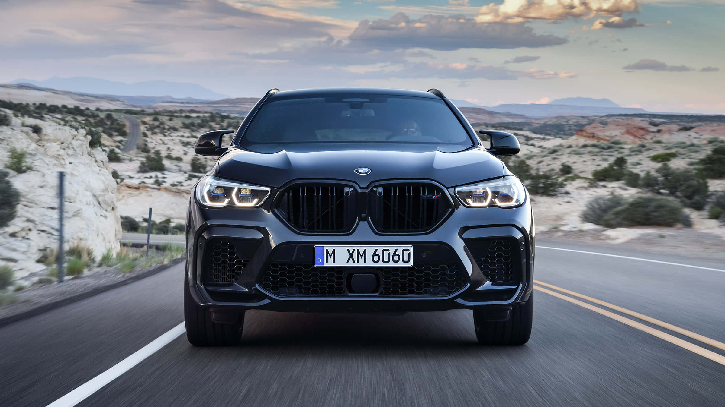 BMW X6 M 桌布 壁紙 — 一輛時尚的黑色BMW XM 6060 SUV在沙漠公路上向前行駛，天空多雲，遠處有山脈。