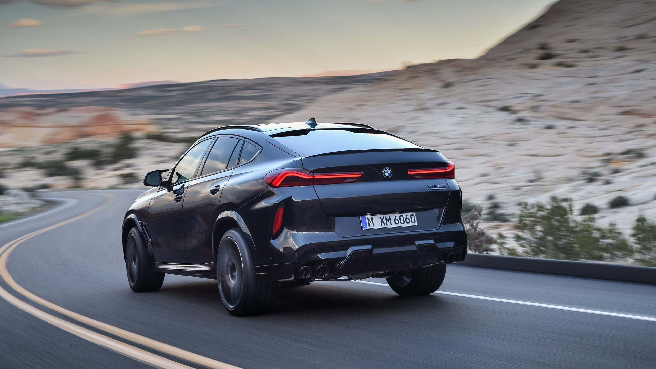 BMW X6 M 桌布 壁紙 — 一輛時尚的黑色BMW XM SUV在日落時分沿著蜿蜒的沙漠公路行駛，帶有模糊的動態和溫暖的金色光線。