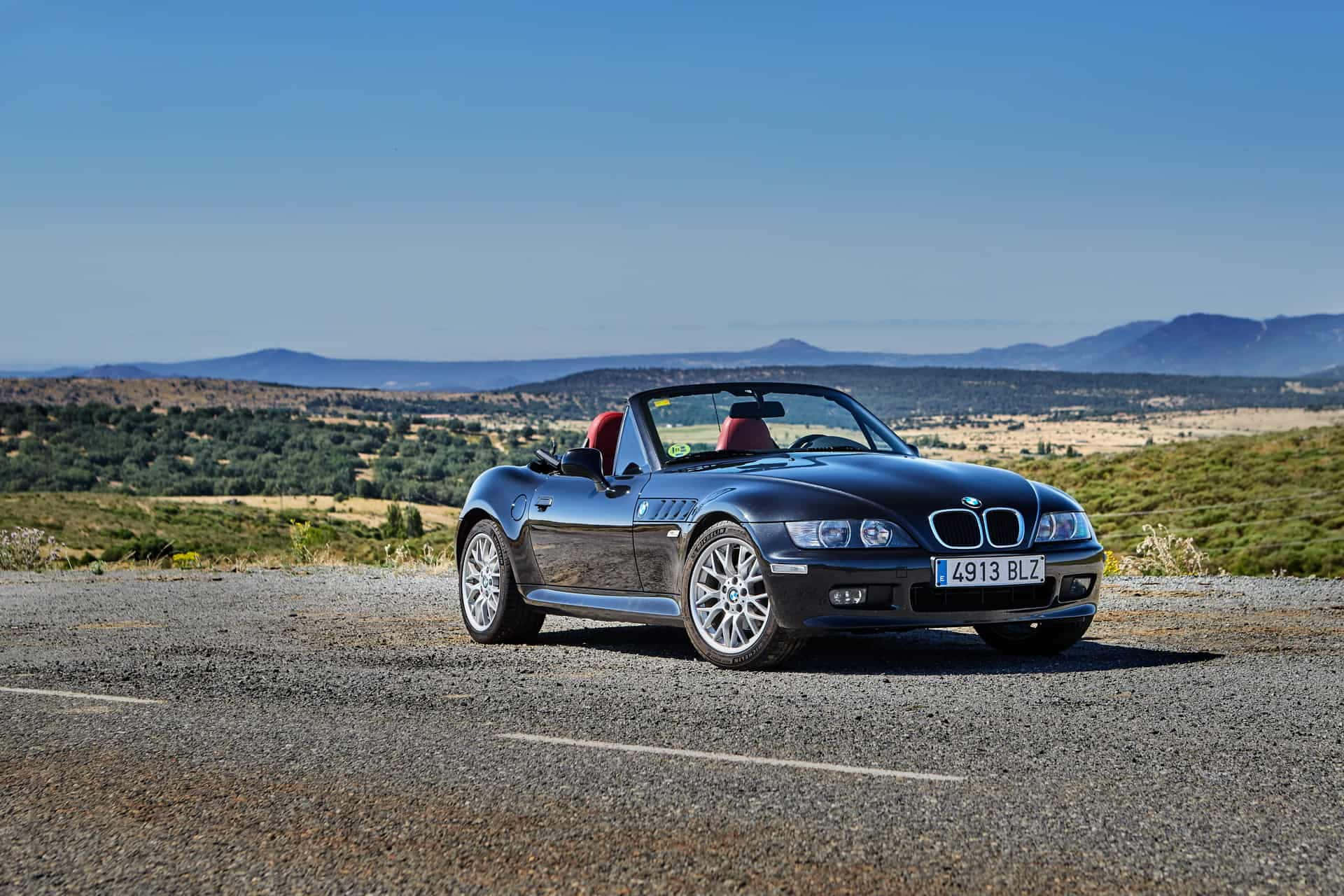 Bmw Z3 桌布 壁紙 — 一輛時尚的黑色BMW Z3敞篷跑車，配紅色座椅，停泊在碎石路上，俯瞰連綿起伏的山丘和遠處的山脈，天空清澈湛藍。
