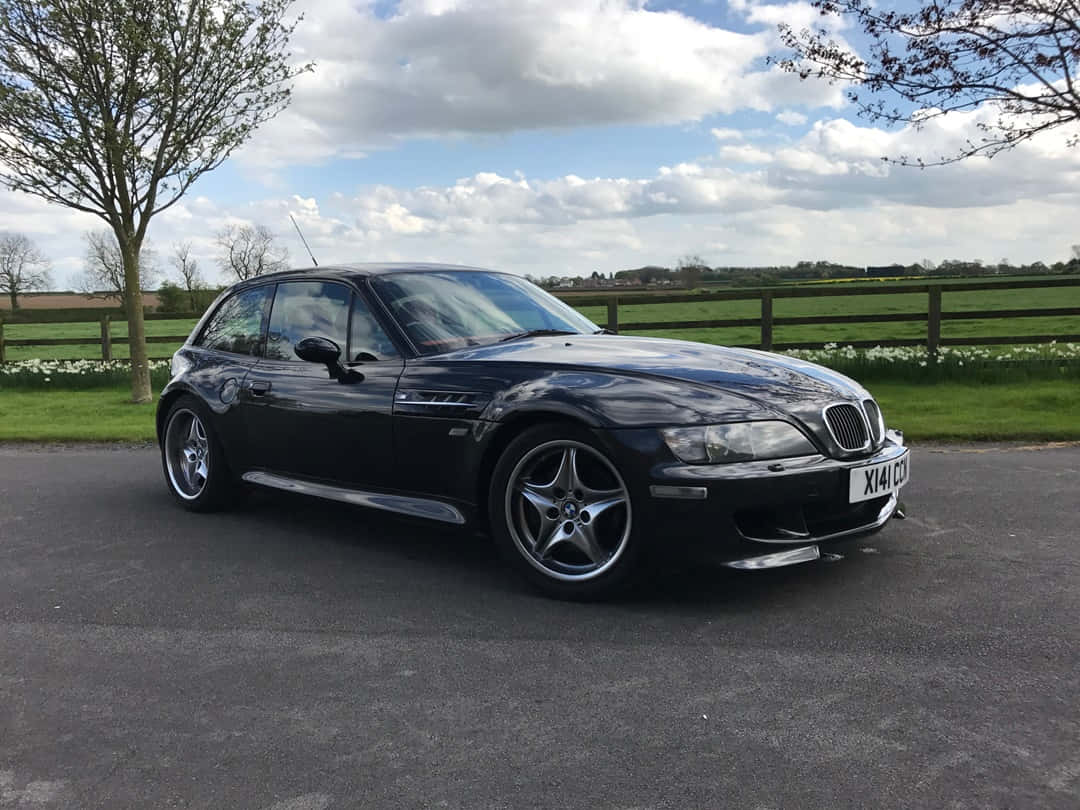 Bmw Z3 桌布 壁紙 — 一輛時尚的黑色BMW Z3跑車停放在鋪砌的車道上，背景是綠色的田野和盛開的白花，天空部分多雲。