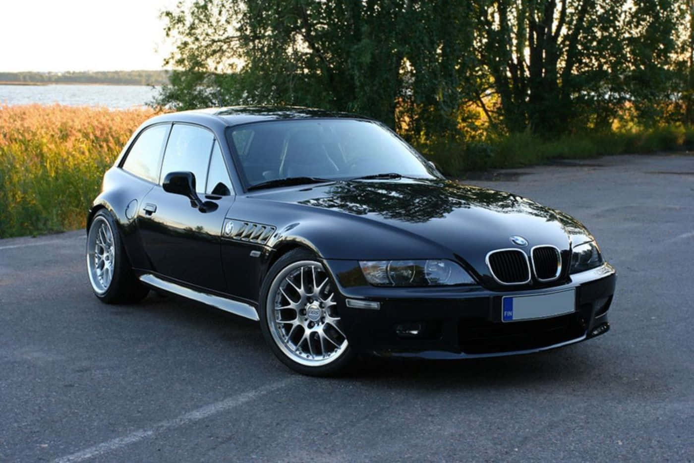 Bmw Z3 桌布 壁紙 — 一輛時尚的黑色BMW Z3跑車在黃金時段停泊在近水邊的高草和樹木旁的瀝青路上。