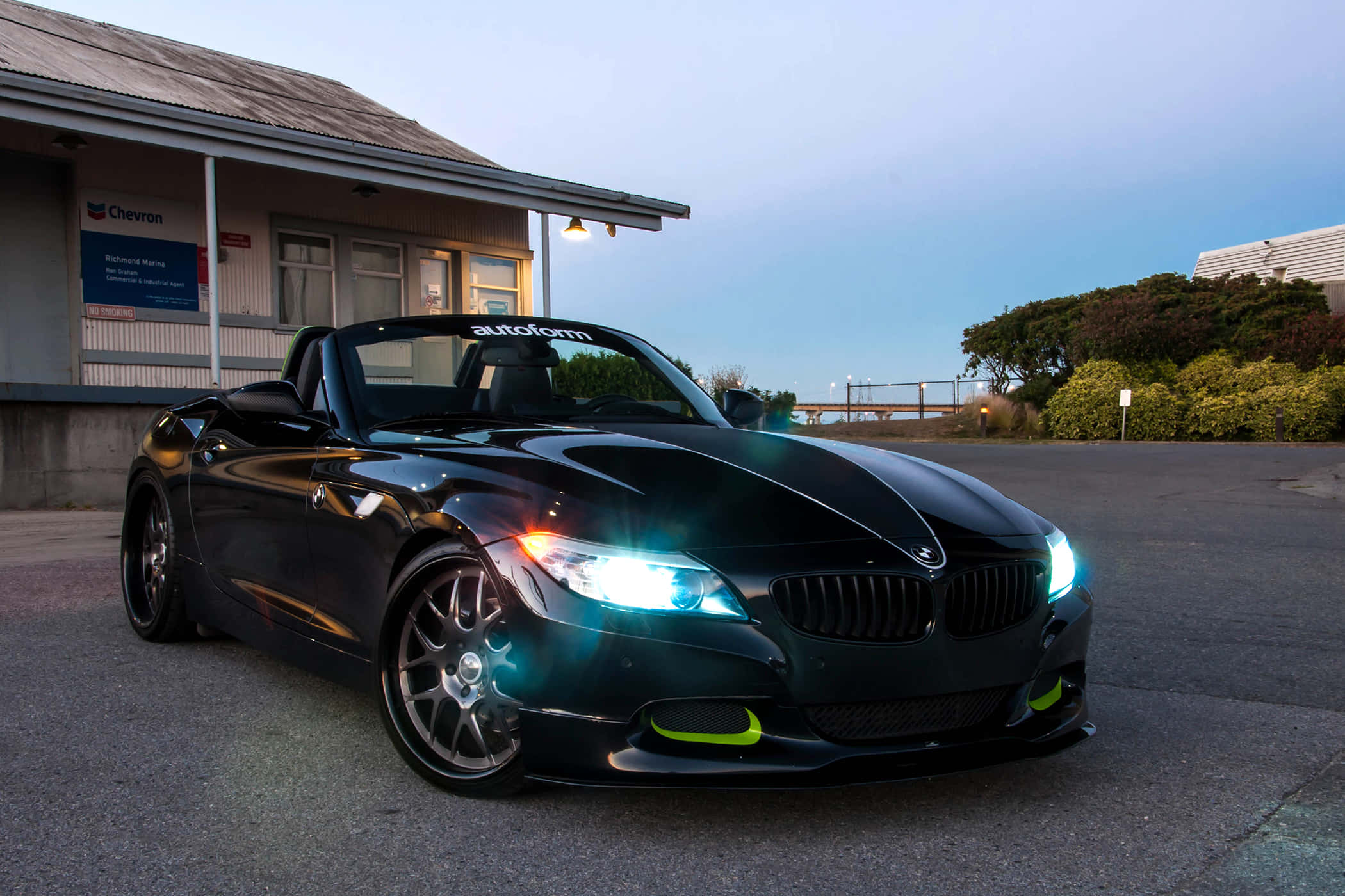 Bmw Z4 桌布 壁紙 — 一輛時尚的黑色BMW Z4敞篷跑車，車頭燈閃爍，停泊在雪佛龍加油站前，正值黃昏。