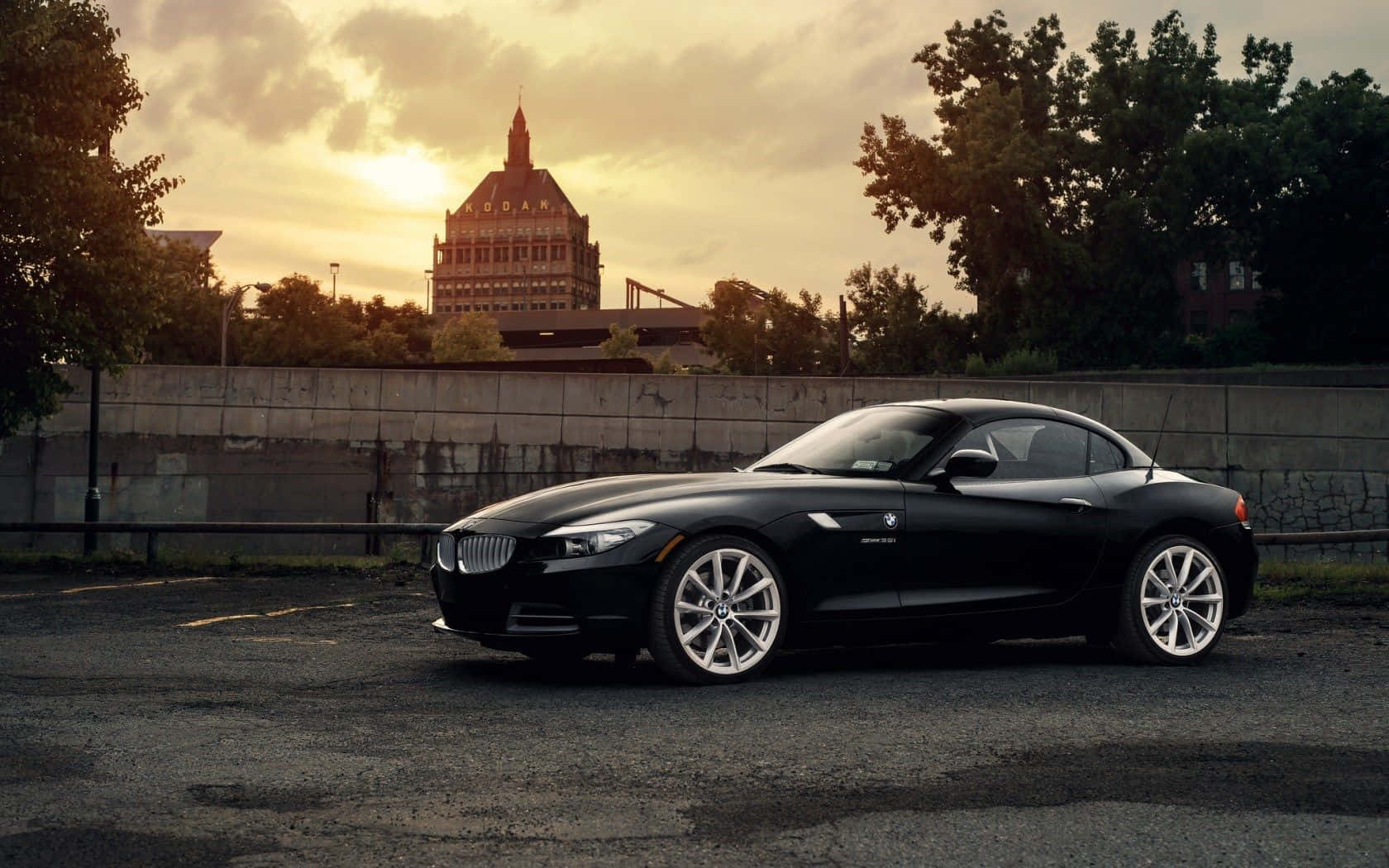 Bmw Z4 桌布 壁紙 — 一輛時尚的黑色BMW Z4跑車停放在日落時分的城市空地，背景有歷史建築和樹木。
