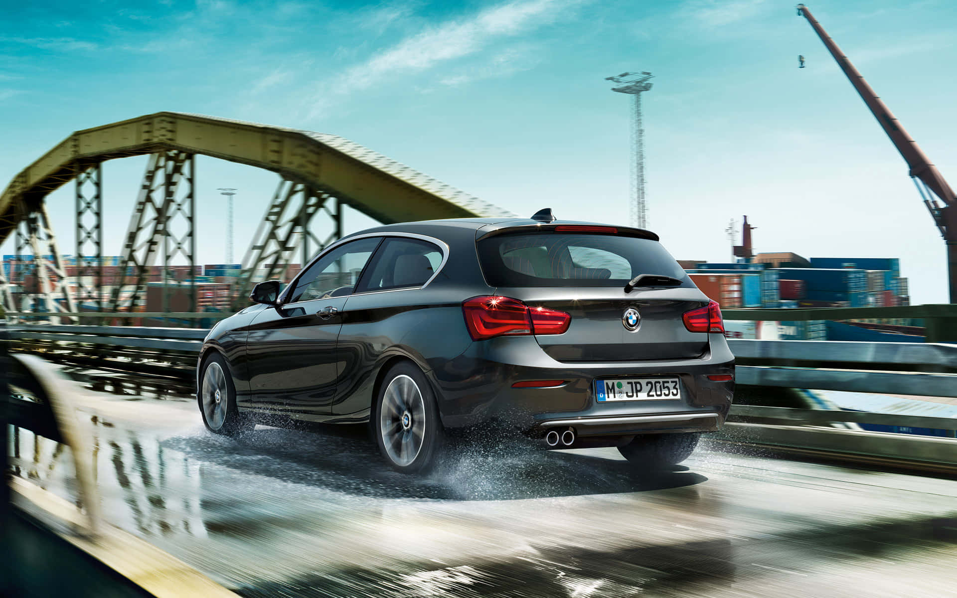 Bmw 1 Series 桌布 壁紙 — 一輛時尚的黑色BMW掀背車在濕滑的橋上飛馳，背景是貨運集裝箱和起重機，天空湛藍清澈。