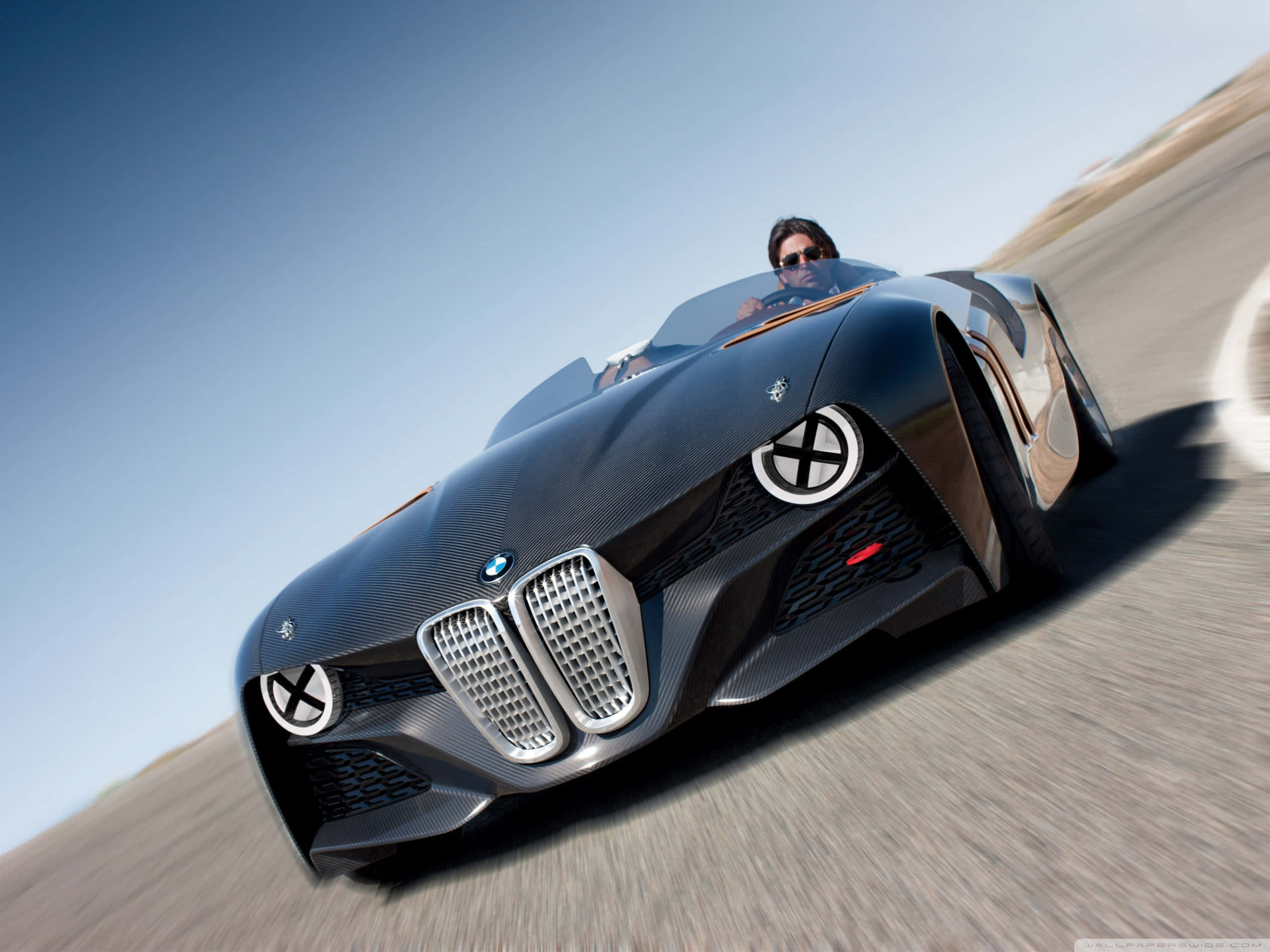 Bmw 328 桌布 壁紙 — 一輛時尚的黑色BMW概念車在晴朗藍天下沿沙漠道路疾馳，司機戴著太陽眼鏡。