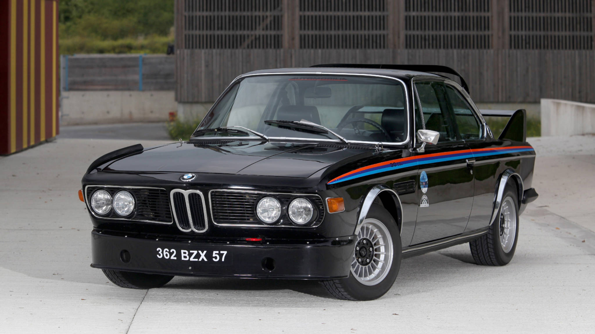 classic bmw 桌布 壁紙 — 一輛時尚的黑色BMW賽車，帶有藍色、紅色和白色條紋，停在木柵欄附近的混凝土上。