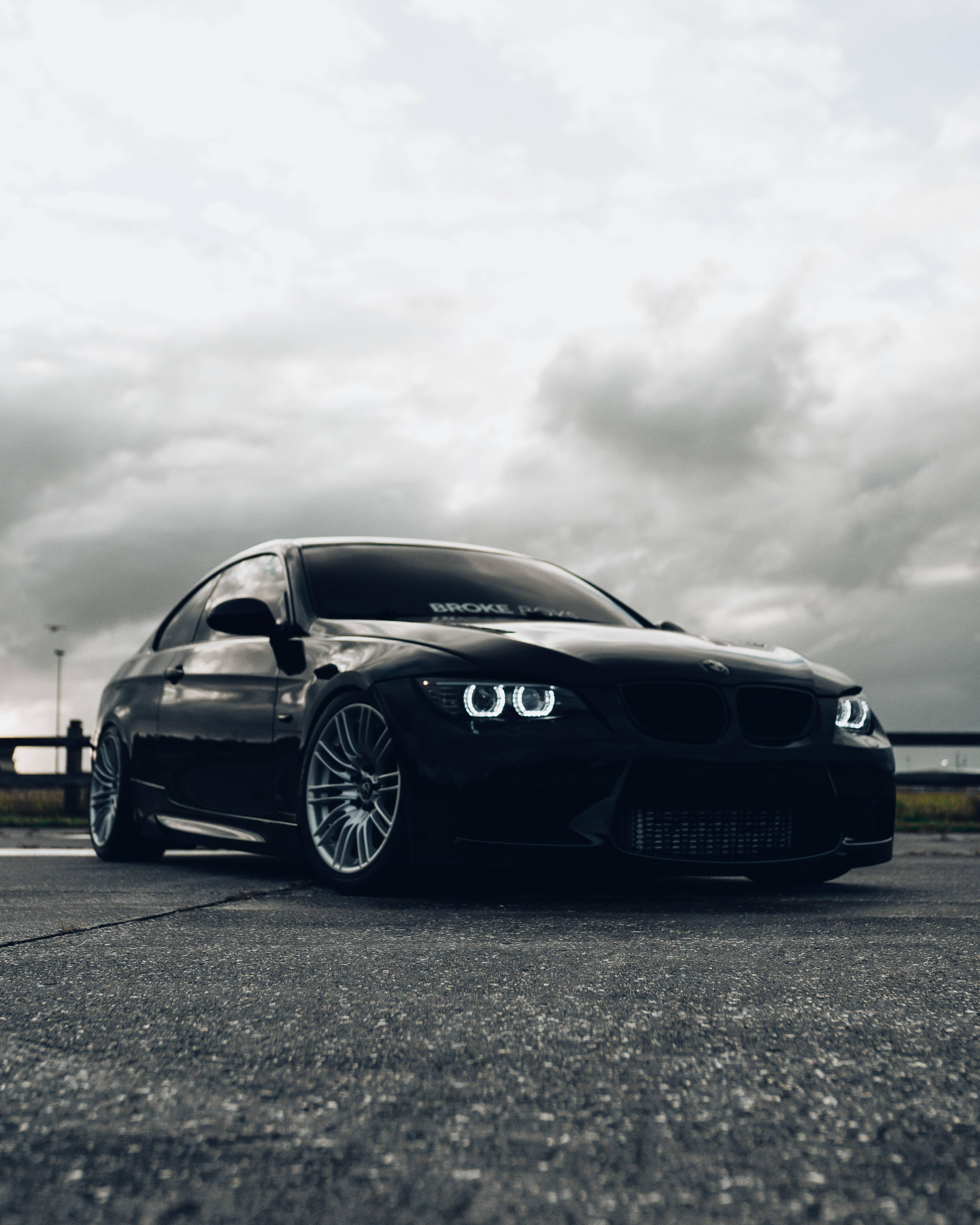 Black Bmw 桌布 壁紙 — 一輛時尚的黑色BMW跑車，配備發光的車頭燈，停在多雲天空下的瀝青路面上。