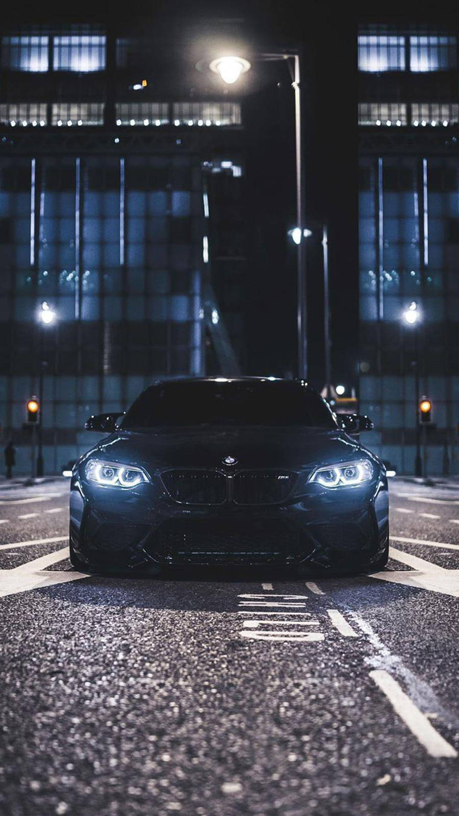 m 桌布 壁紙 — 一輛時尚的黑色BMW跑車在夜間濕漉漉的城市街道上，在街燈下閃耀，背後是反光的玻璃建築。