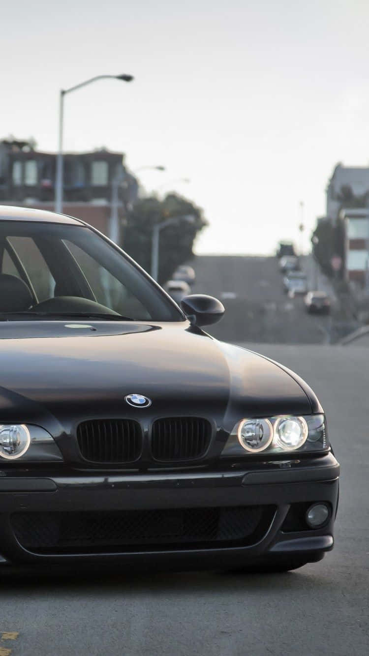 Bmw iPhone 桌布 壁紙 — 一輛時尚的黑色BMW跑車在柔和的日光下閃耀，停在一條寧靜的城市街道上，背景是模糊的建築物和街燈。