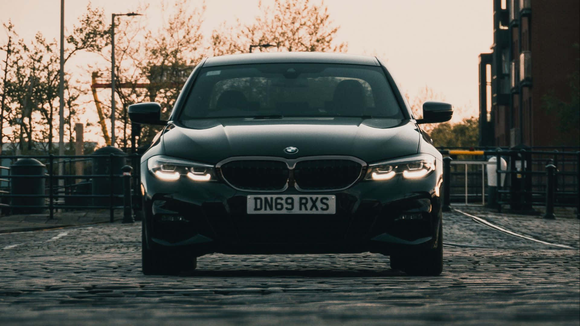 Car Images HD 桌布 壁紙 — 一輛時尚的黑色BMW轎車，車頭燈發亮，在黃金時段面向鵝卵石街道的前方。