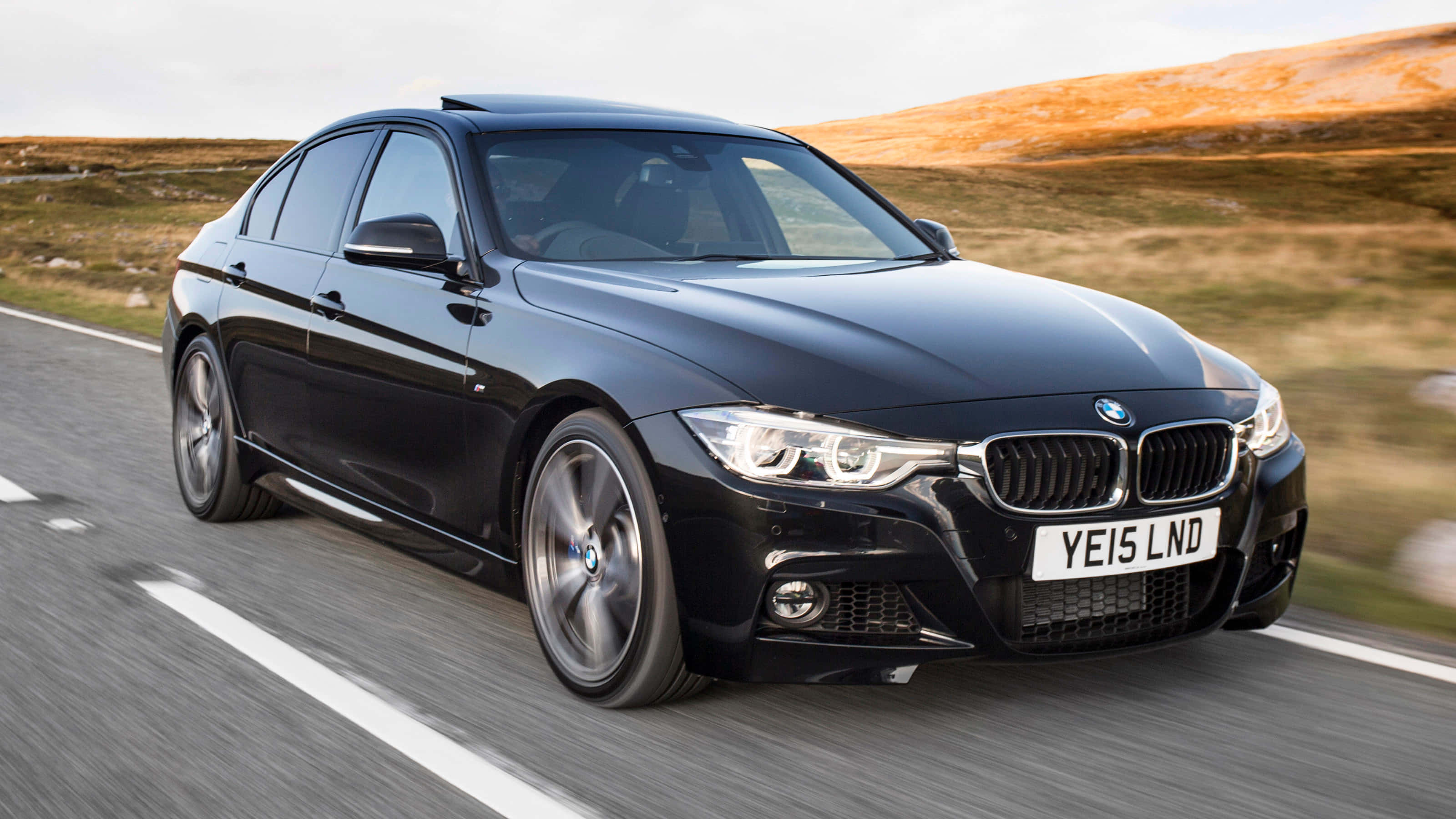 Bmw 3 Series 桌布 壁紙 — 一輛時尚的黑色BMW轎車在晴朗天空下，沿著蜿蜒的道路穿過起伏的金色山丘飛馳。
